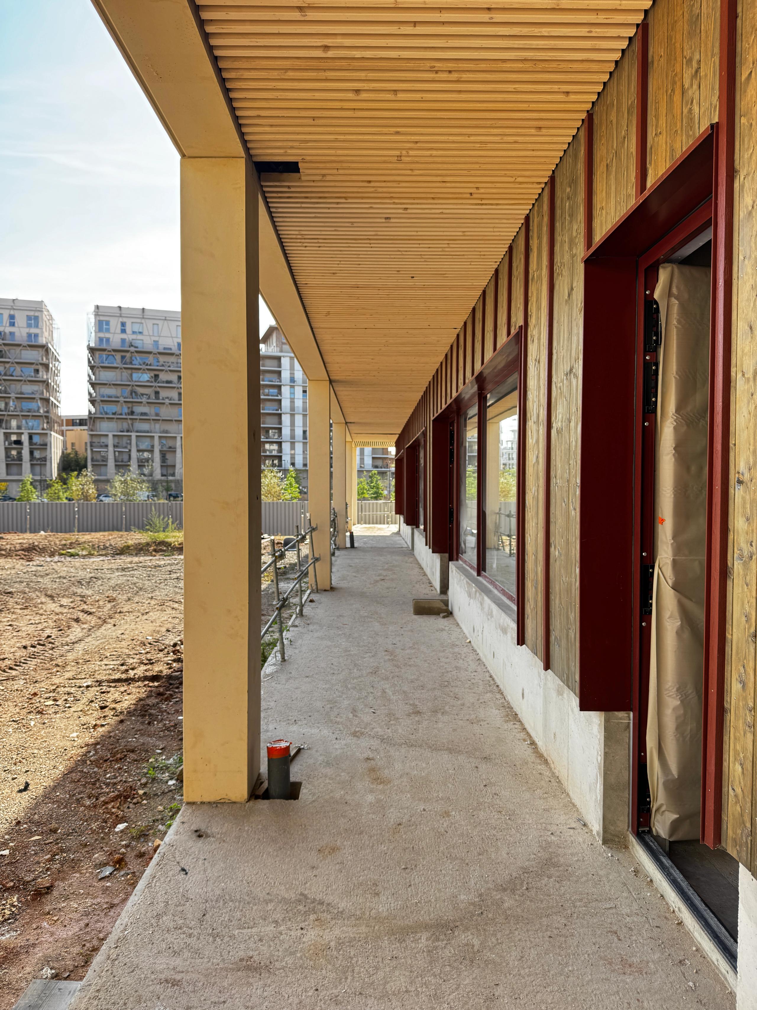 Coursive couverte du groupe scolaire Frida Kahlo à Bordeaux : poteaux et sous-face en bois, façade bardée et encadrements métalliques rouges.