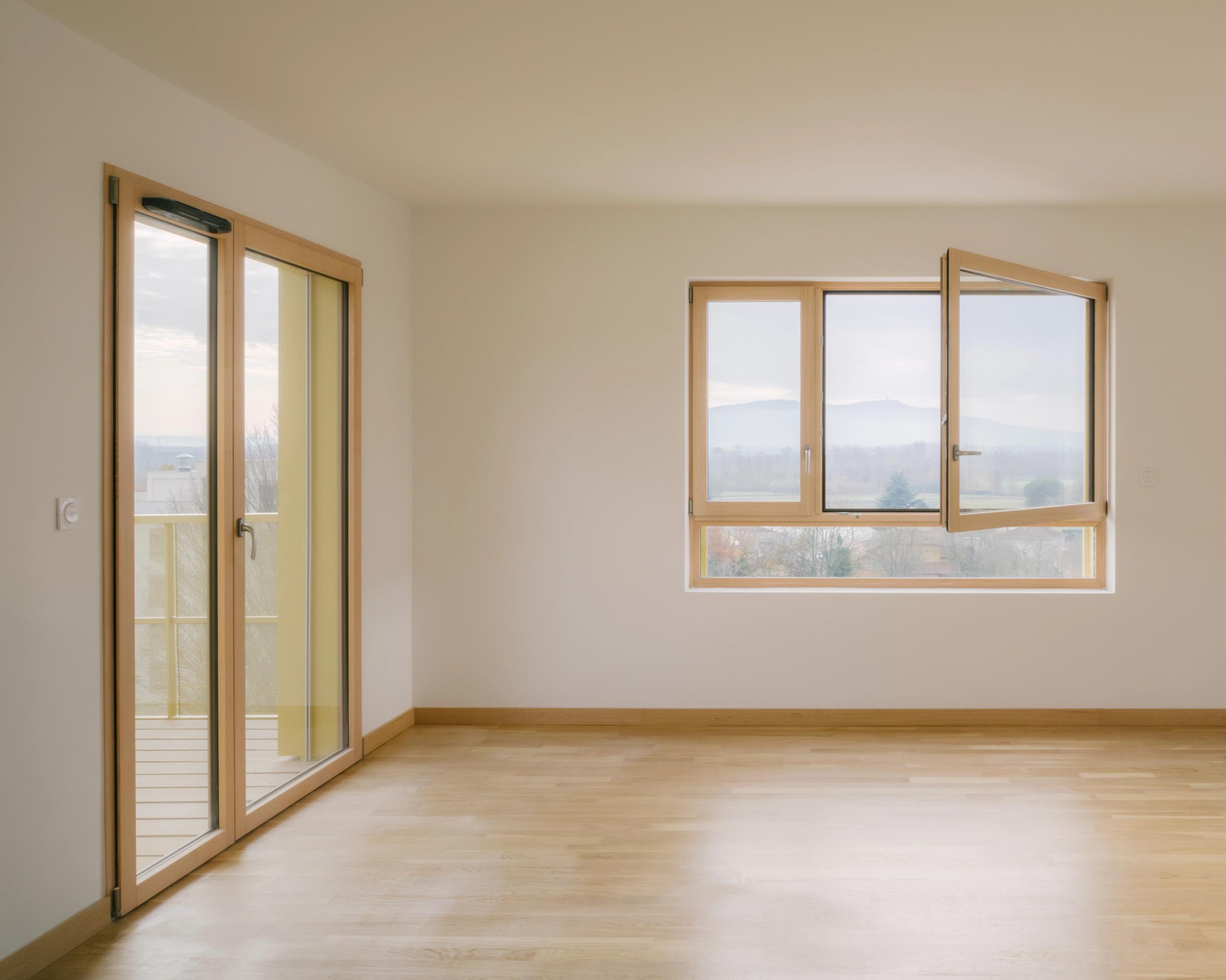 Intérieur d’un logement des 84 logements à Trévoux, pièce vide avec menuiseries bois-aluminium, grande baie sur balcon et fenêtre cadrant le paysage.