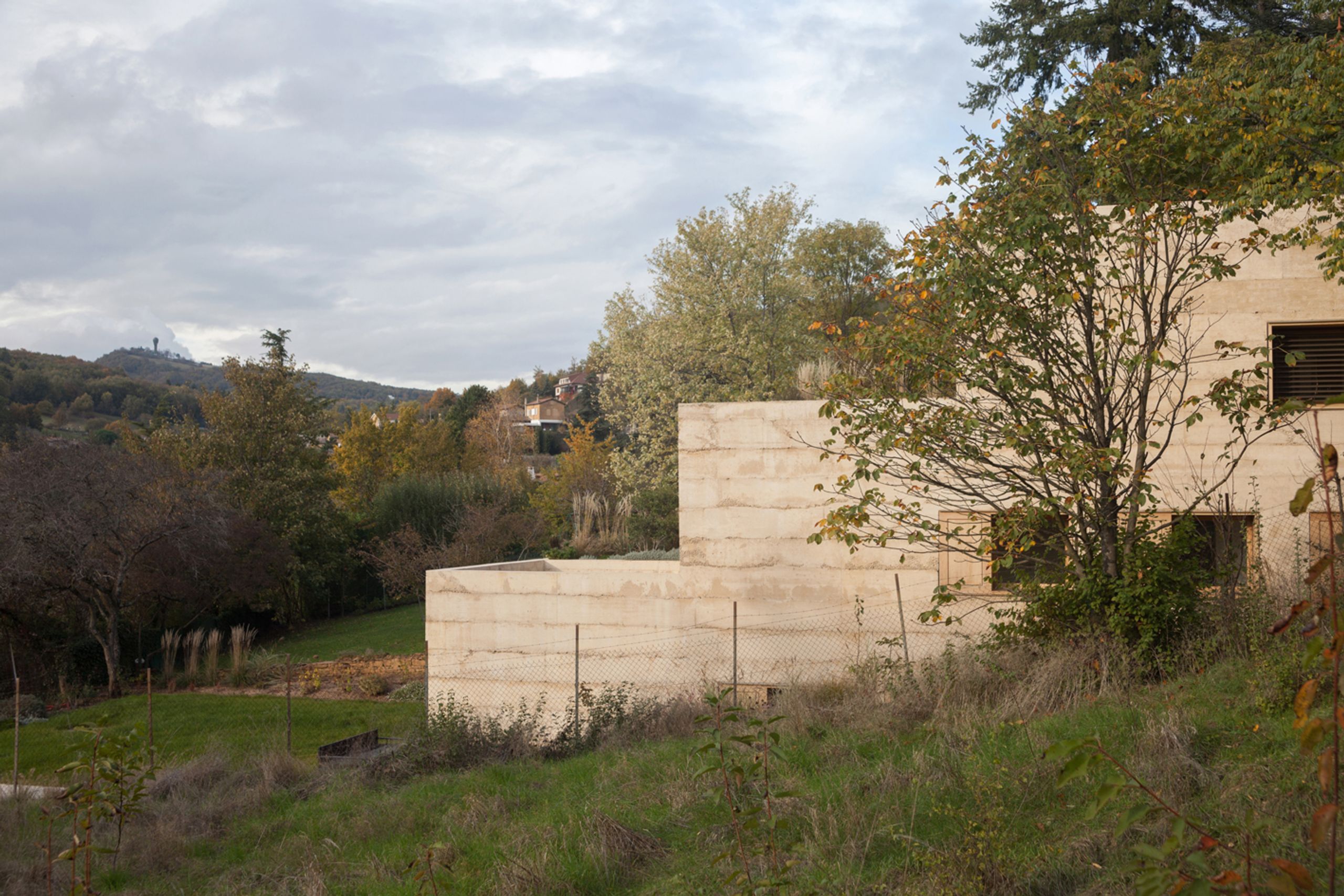 Maison en béton teinté en strates, encastrée dans un vallon à Saint-Cyr-au-Mont-d’Or, volumes en terrasses ouverts sur le paysage boisé environnant.