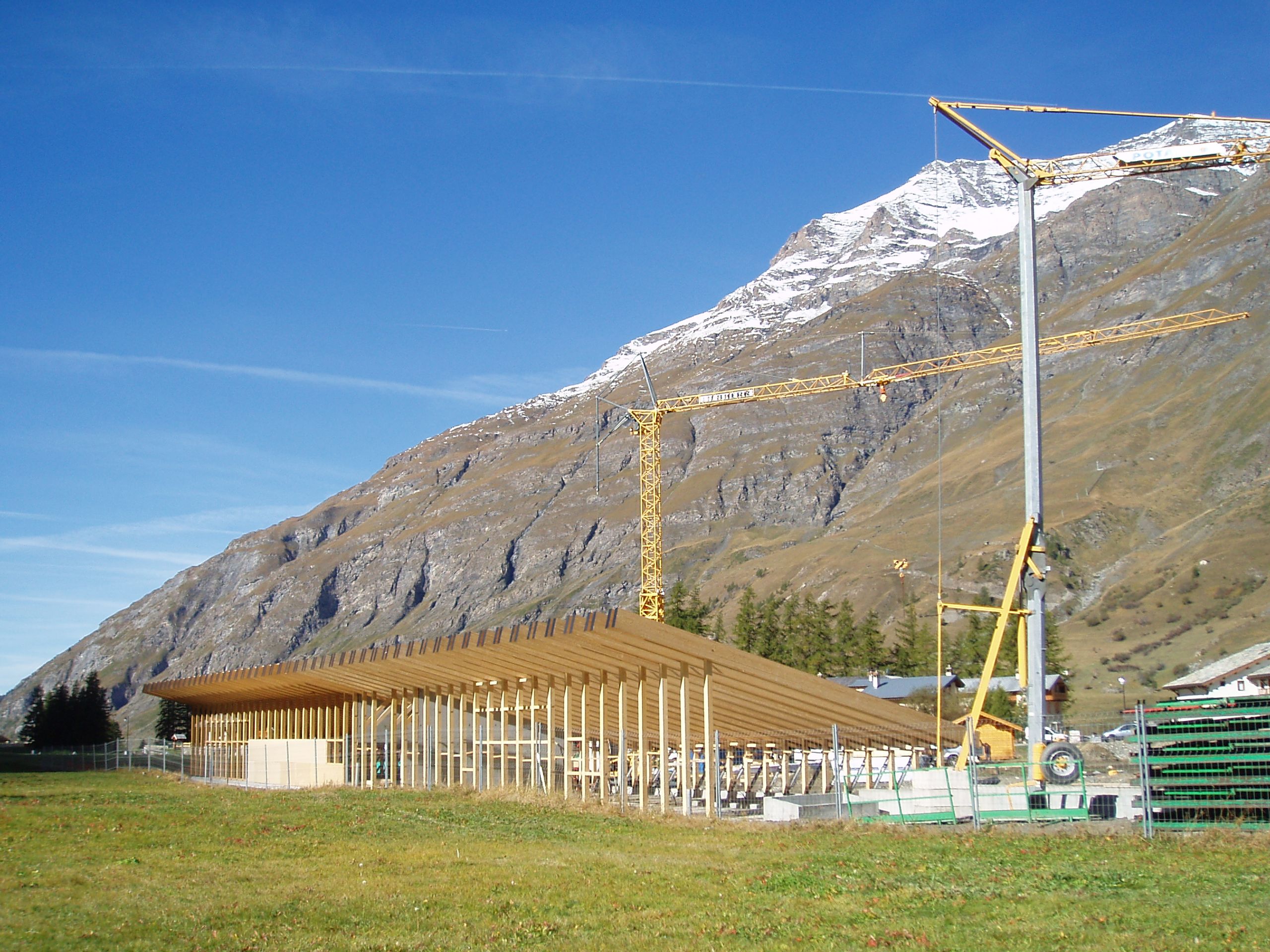 Chantier de l’espace nordique de Bessans : volume bas en bois à toiture inclinée, portiques réguliers face aux pistes, inséré au pied du massif alpin.