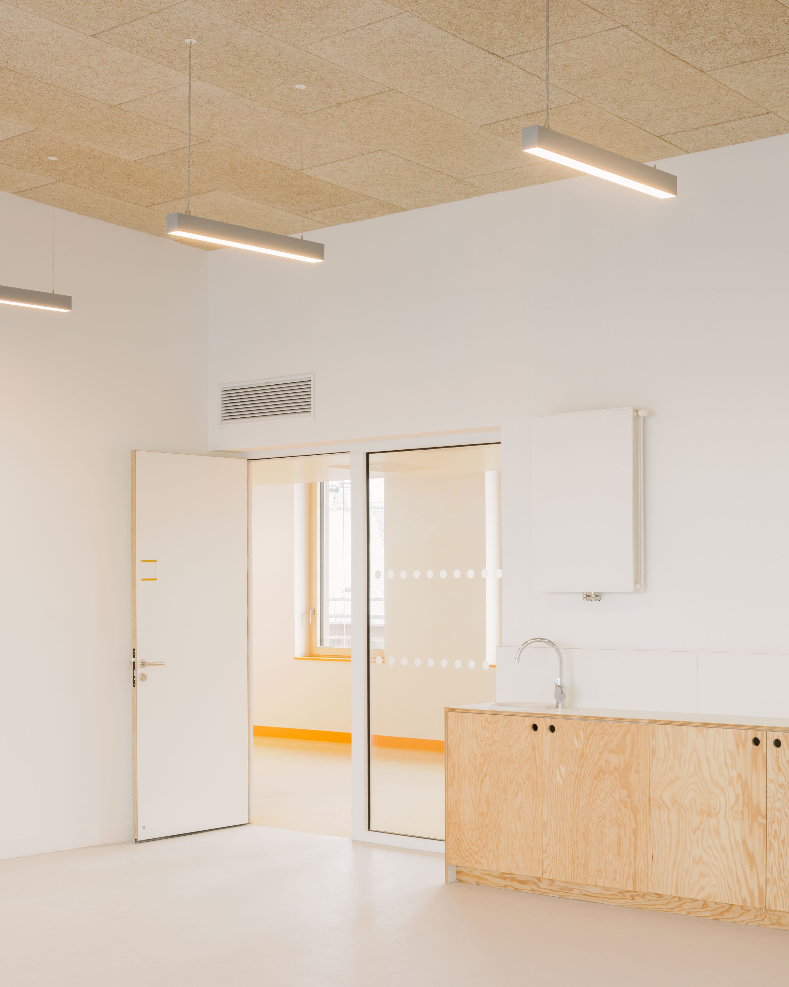 Intérieur lumineux des groupes scolaires Jacqueline Auriol et Jean Jaurès au Bourget, avec plafond en dalles minérales, menuiseries bois et cloison vitrée.