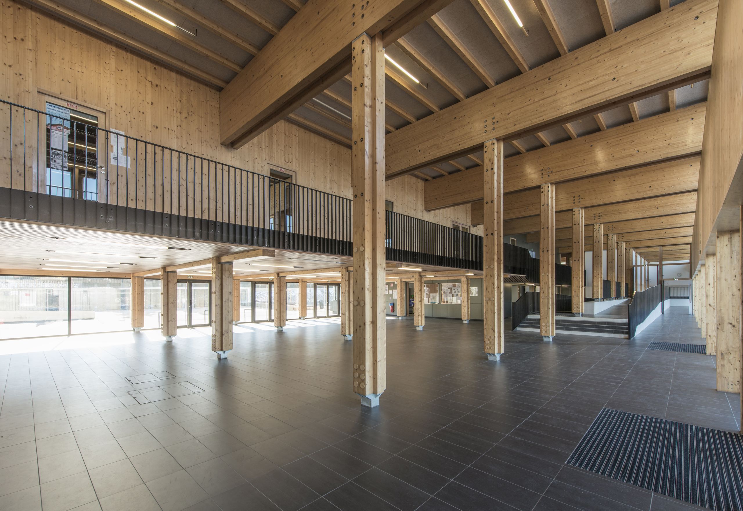 Grande galerie traversante du collège Pierre-Joseph Proudhon à Besançon, structure poteau-poutre bois apparente, mezzanine et rampes noires sur sol carrelé sombre.