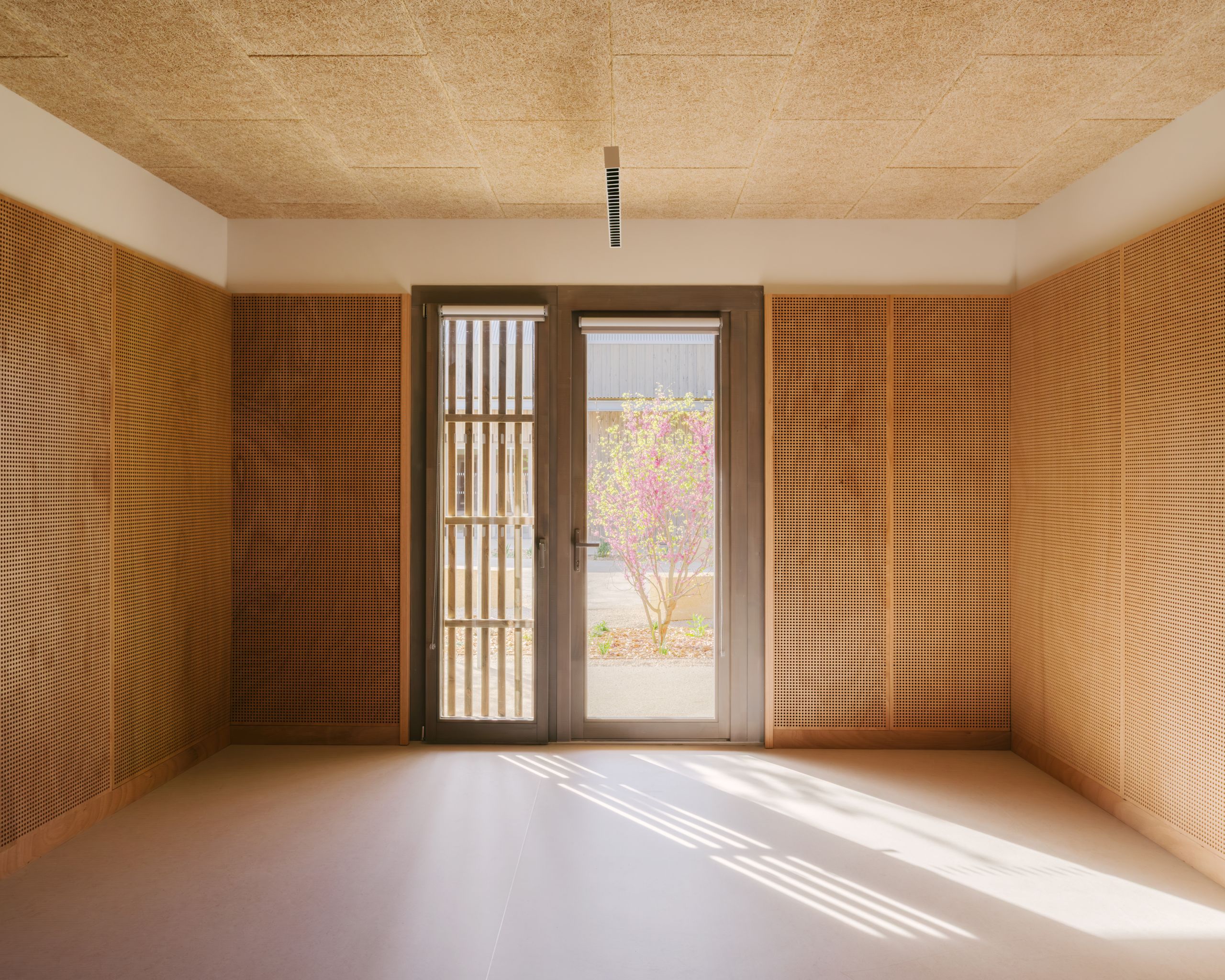 Intérieur du pôle culture et jeunesse à Vaison-la-Romaine : salle aux parois bois ajouré et plafond absorbant, ouverte par une porte vitrée sur le jardin.