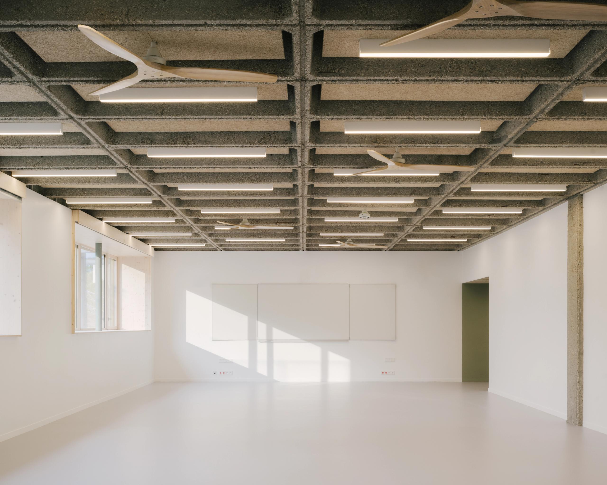 Salle de classe restructurée du collège Maryse Bastié à Dole, plafond à caissons béton apparents, éclairage linéaire, parois blanches et ouvertures latérales.
