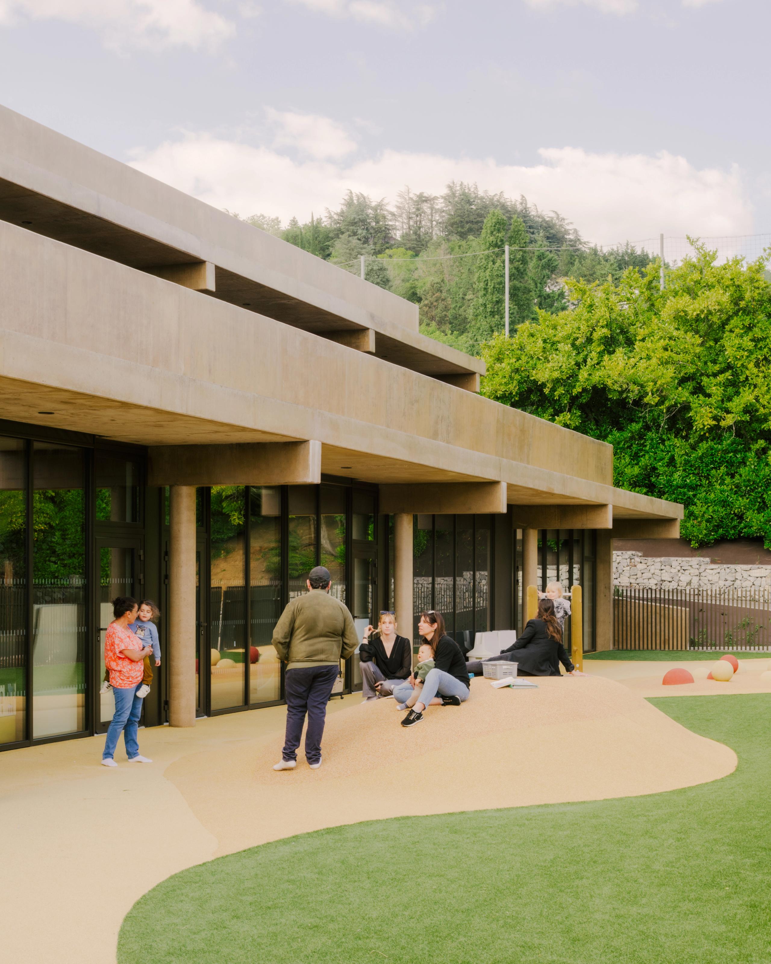 Maison des familles à Limonest : volume en béton teinté en terrasses, larges baies vitrées sur cour paysagée, espaces extérieurs dédiés à la crèche.