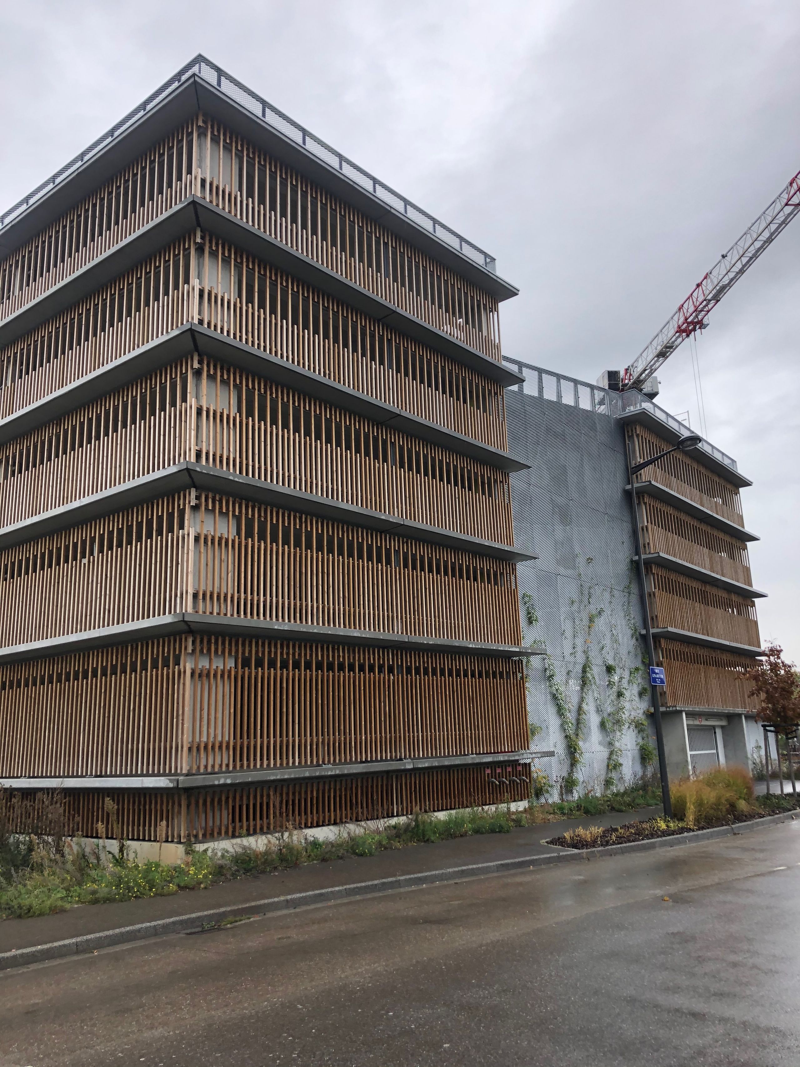 Façade du parking silo Tectoniques à Strasbourg, structure béton et bardage de lames bois verticales, pignon métallique végétalisé sur rue humide.