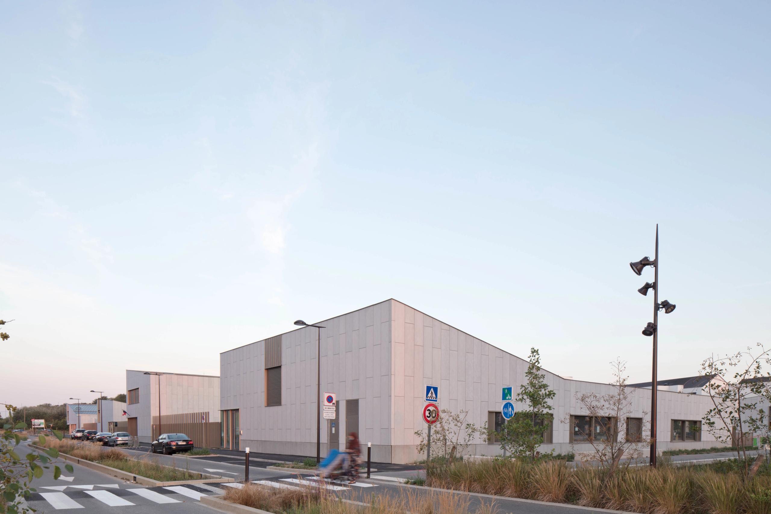 Façades minérales et volumes bas du groupe scolaire Denis Mukwege à Melun, alignés le long de la voirie, percés de baies horizontales et de panneaux bois.