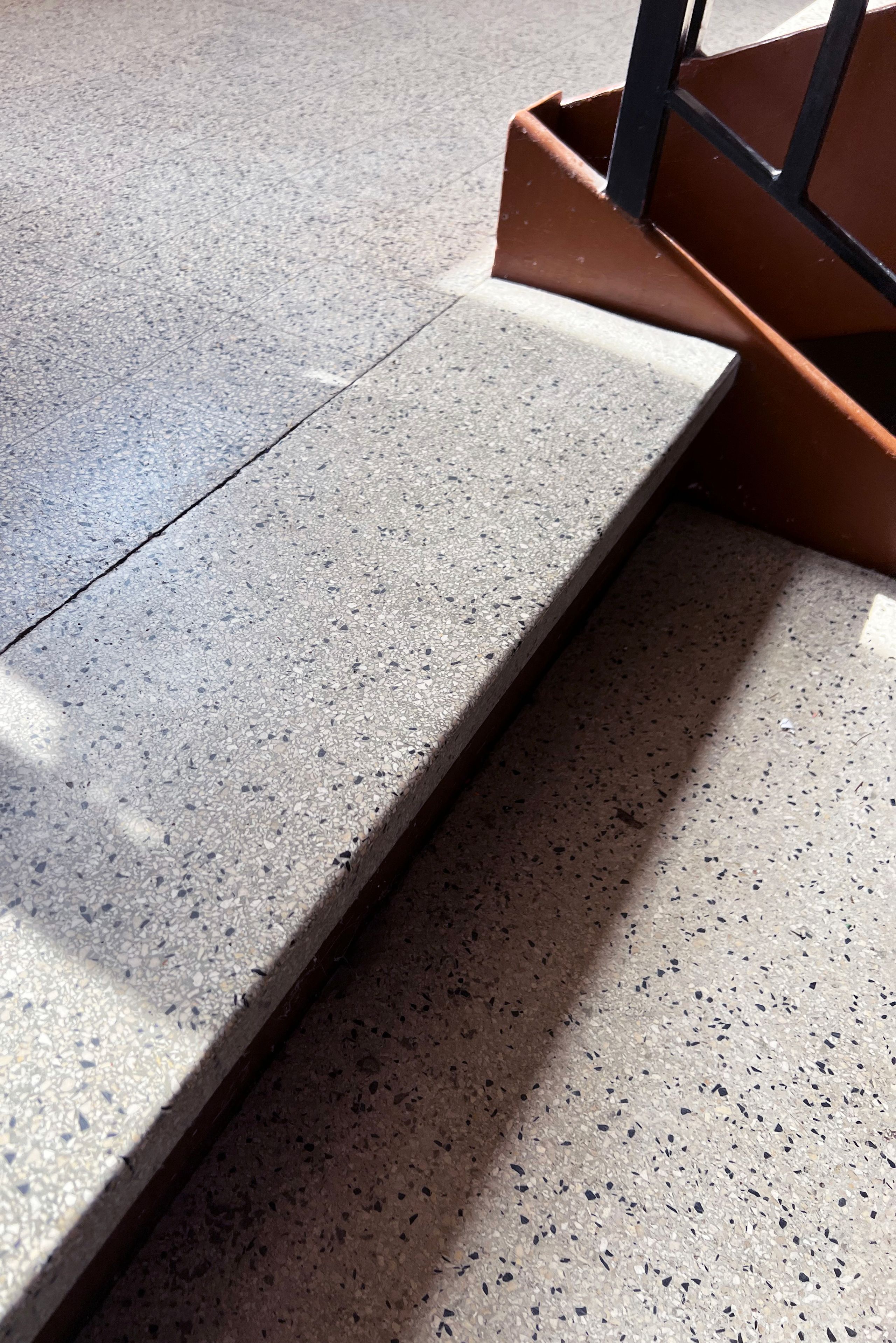 Détail d’escalier intérieur du groupe scolaire Kennedy à Lyon, marche en terrazzo clair, limon métallique brun et garde-corps noir dans la lumière naturelle.