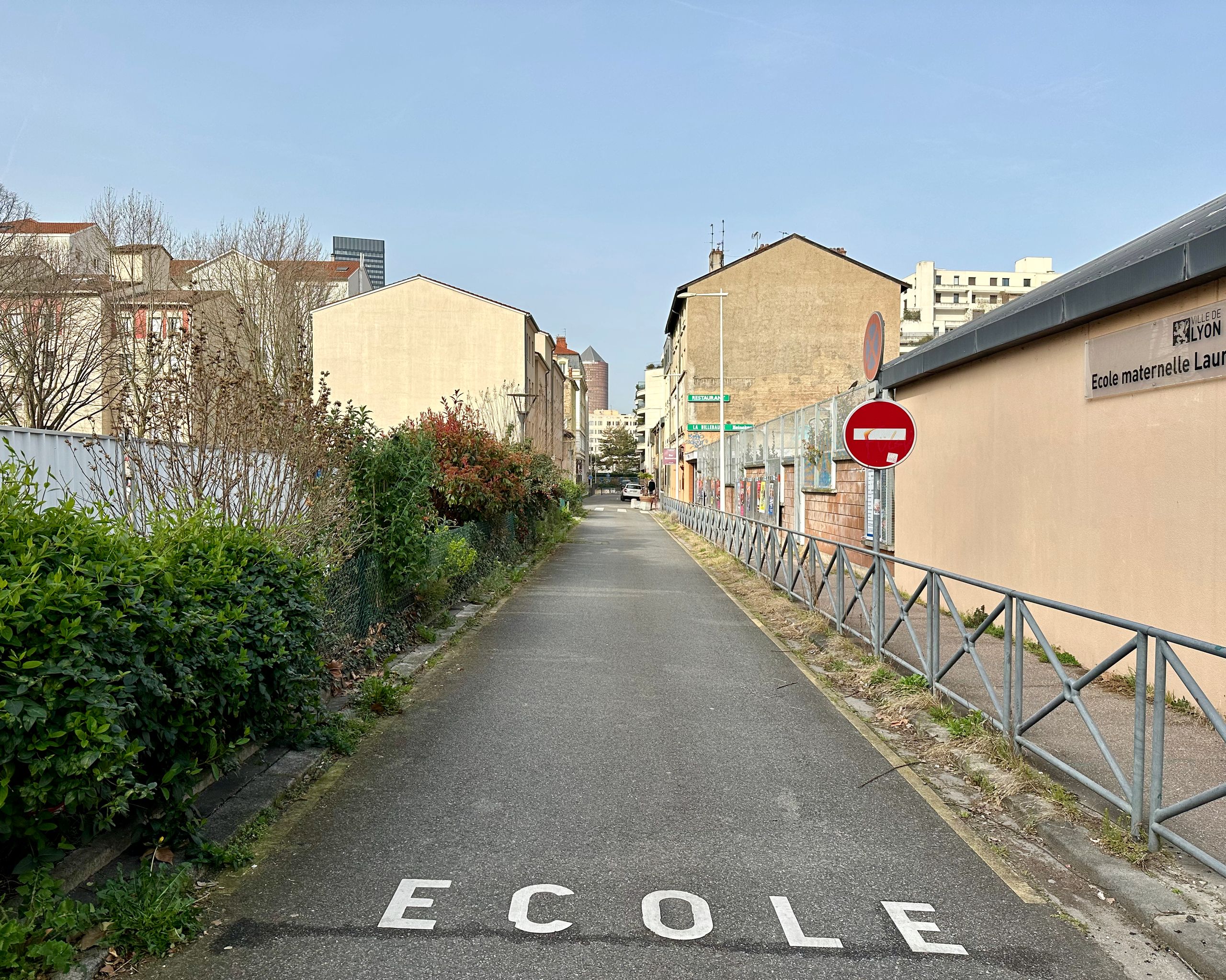Narrow street leading to the Pompidou Mourguet School Complex site in Lyon, framed by existing masonry buildings, school wall and urban vegetation.