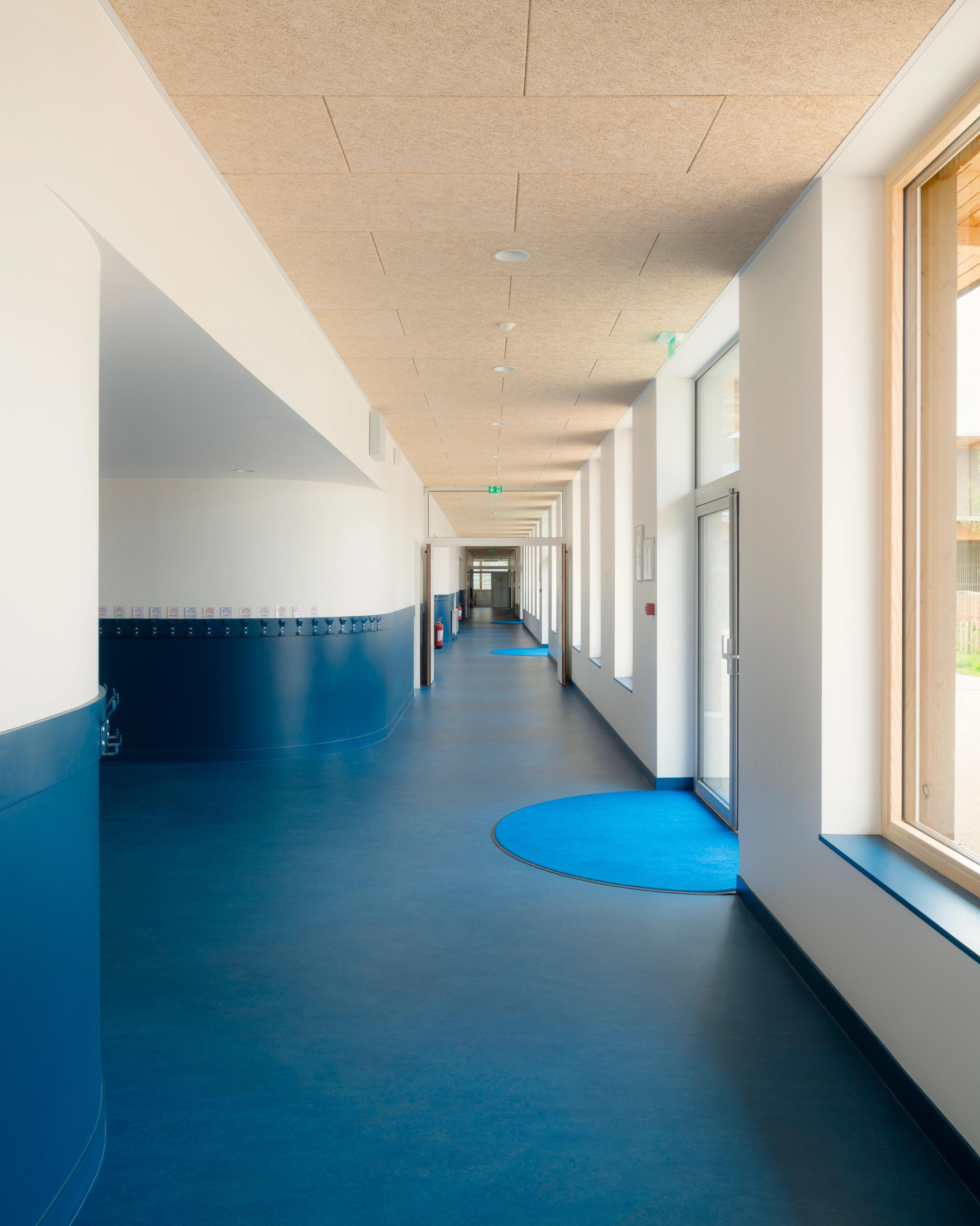 Couloir des groupes scolaires Jacqueline Auriol et Jean Jaurès au Bourget, sol bleu continu, parois blanches, plafond bois et grandes baies vitrées sur cour.