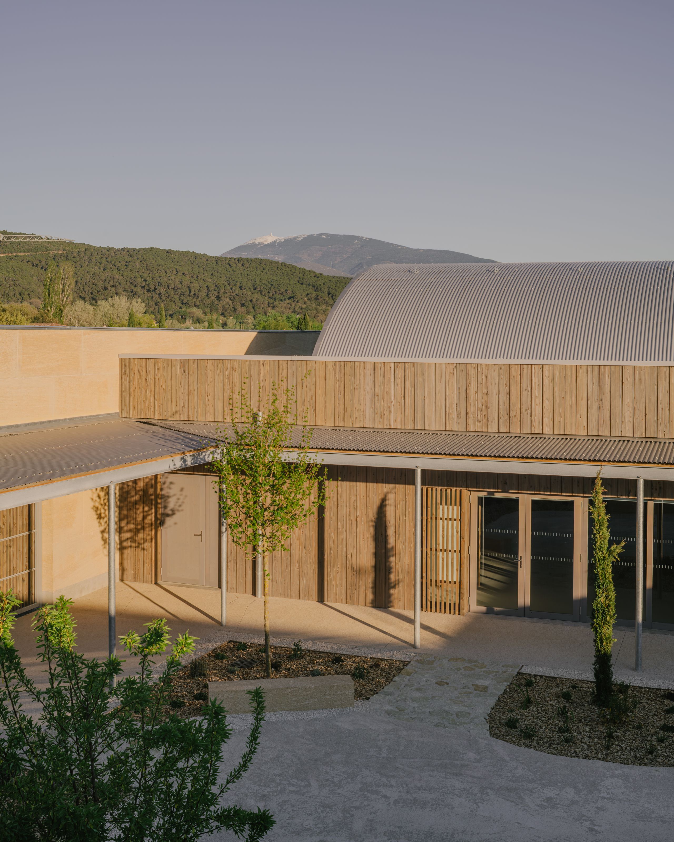 Pôle culture et jeunesse à Vaison-la-Romaine : façade en bois et pierre massive, auvent métallique sur cour minérale plantée, volume courbe de la salle en arrière-plan.