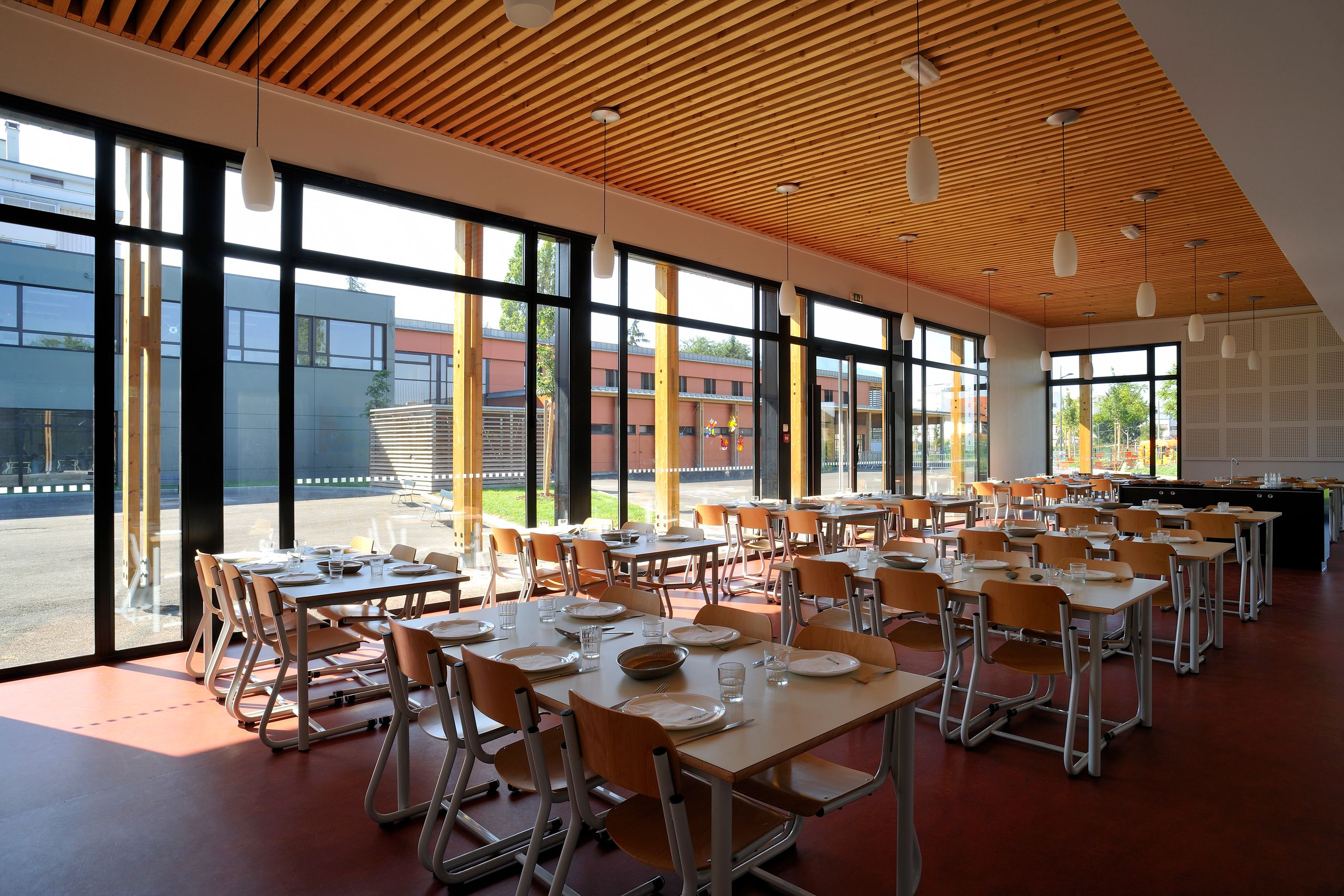 Réfectoire du groupe scolaire Antoine de Saint-Exupéry à Annemasse, vaste volume vitré sur cour, structure bois apparente et plafond à lames, mobilier scolaire aligné.