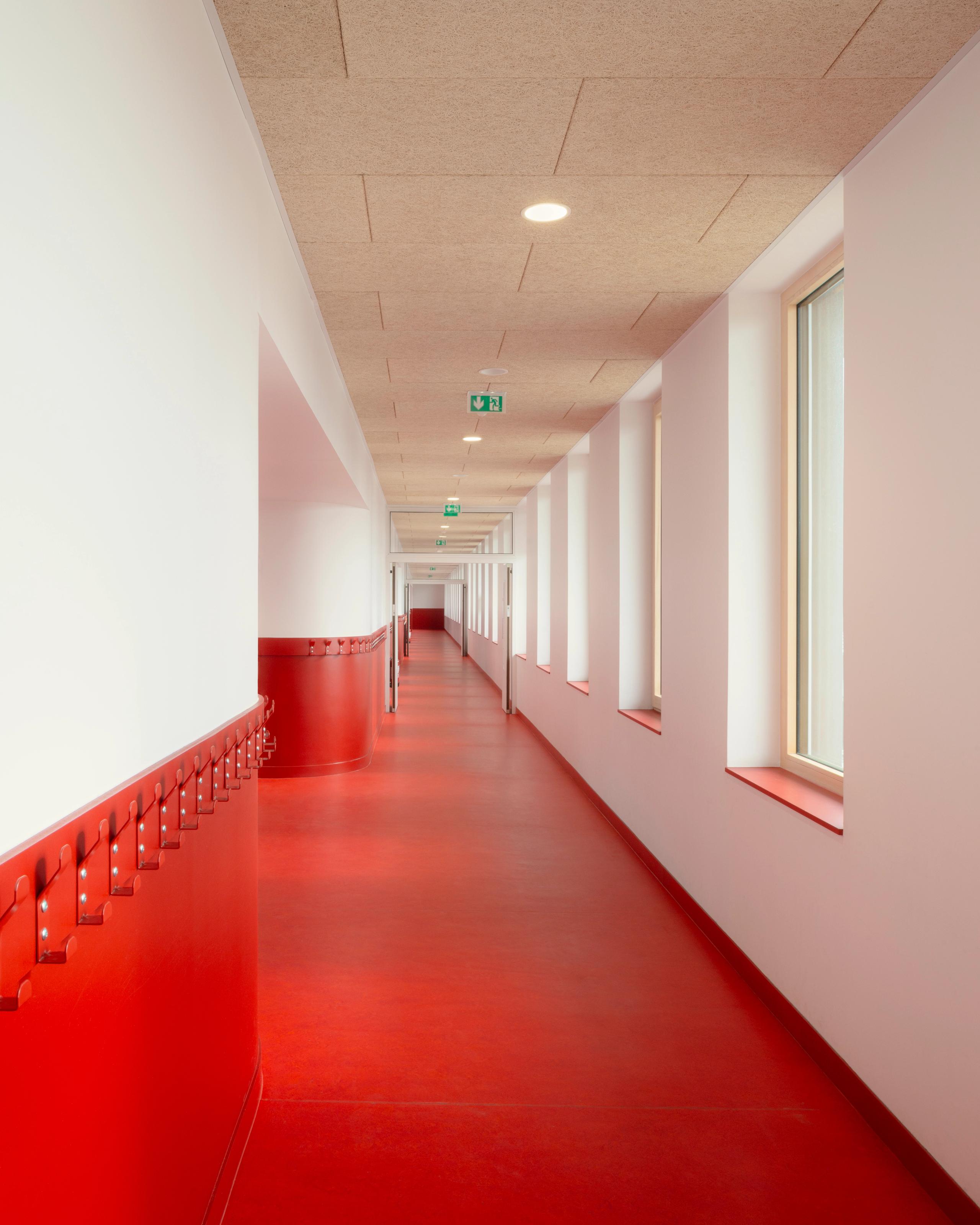 Couloir des groupes scolaires Jacqueline Auriol et Jean Jaurès au Bourget, sol rouge continu, soubassement équipé de patères, baies latérales et plafond acoustique clair.