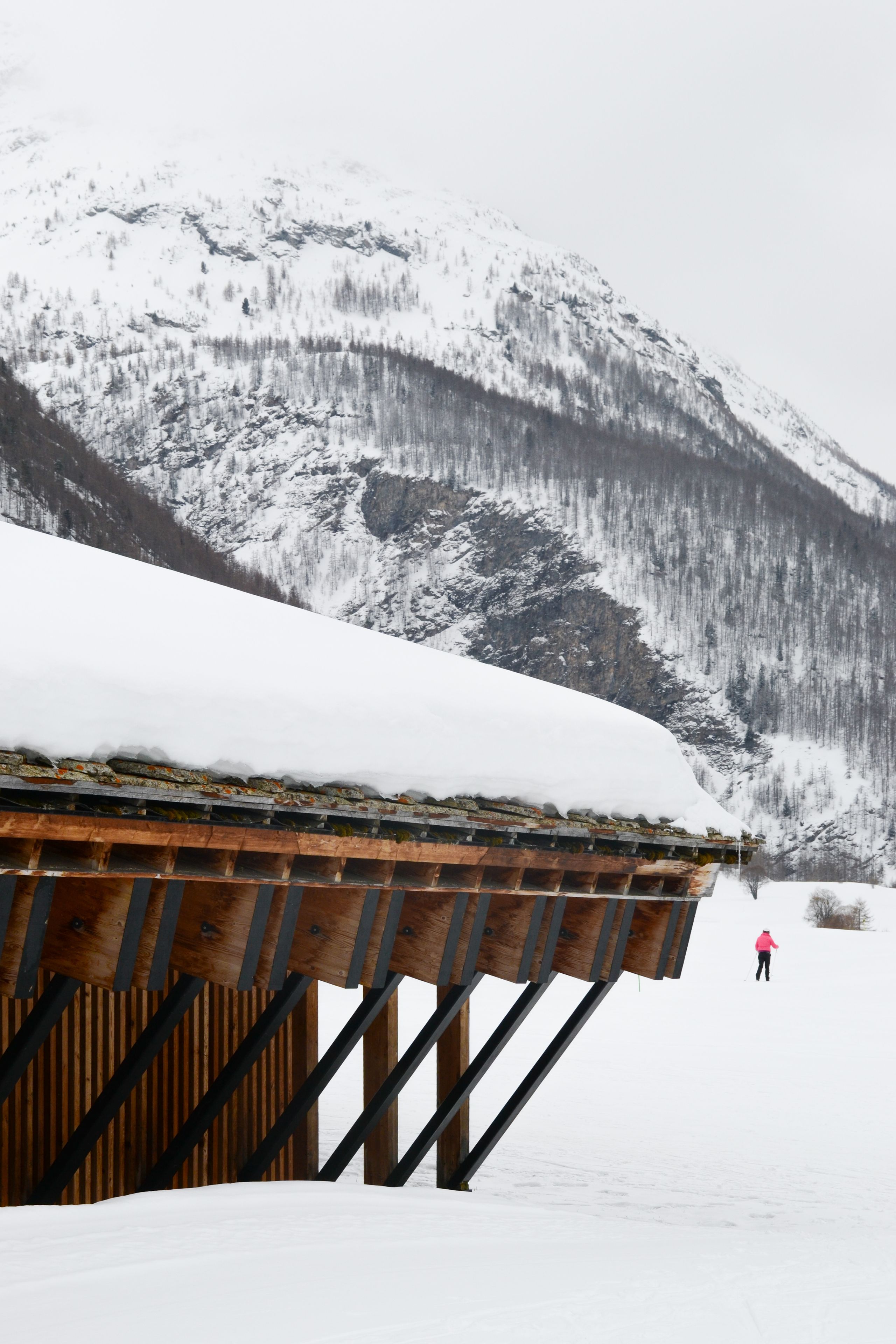 Espace nordique de Bessans : volume en bois sur portiques métalliques inclinés, toiture en lauze enneigée, inséré au pied des pentes boisées de Haute-Maurienne.
