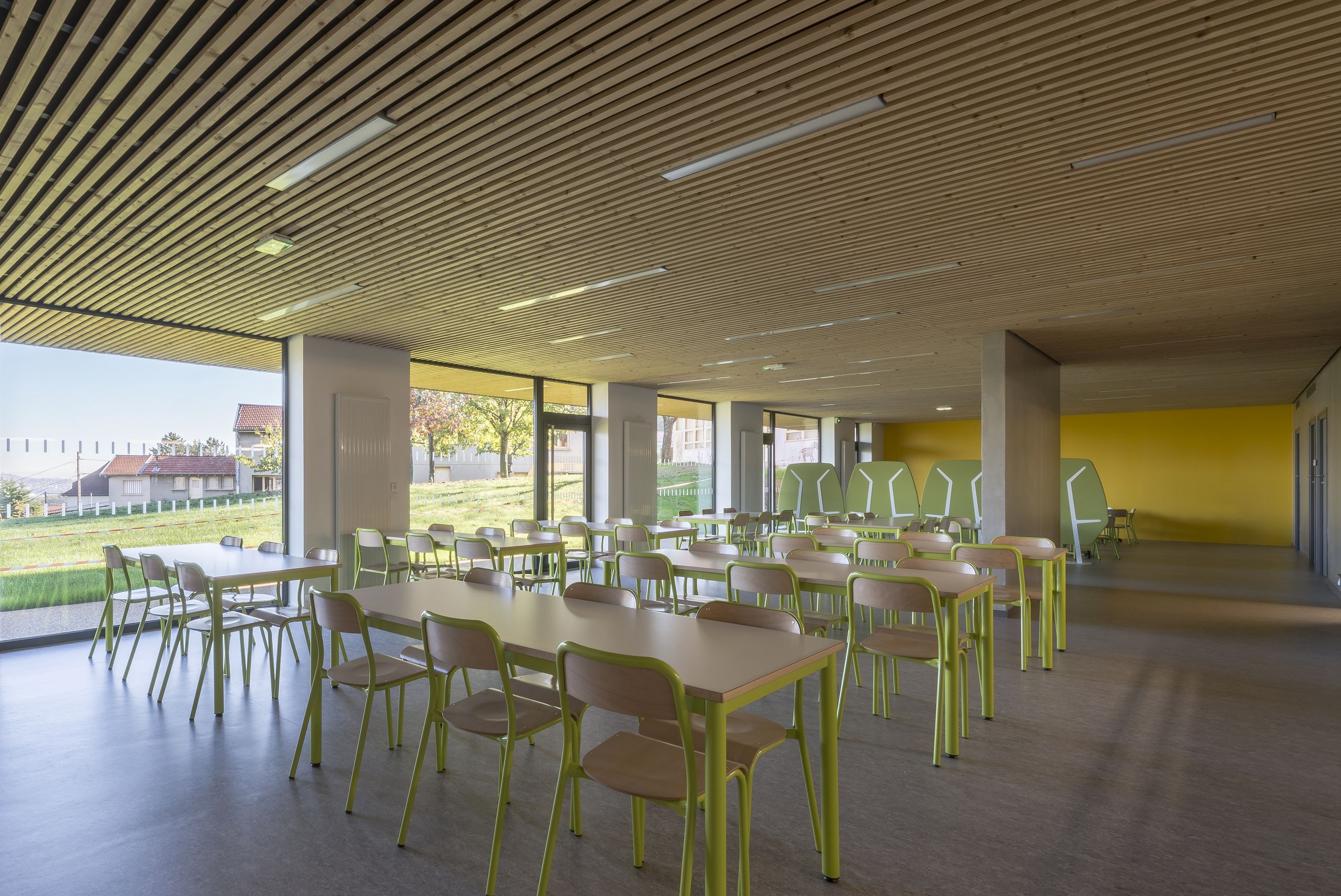 Intérieur du centre de loisirs de Givors : vaste réfectoire vitré sur le paysage, plafond en lames de bois, mobilier vert clair modulable pour les enfants.