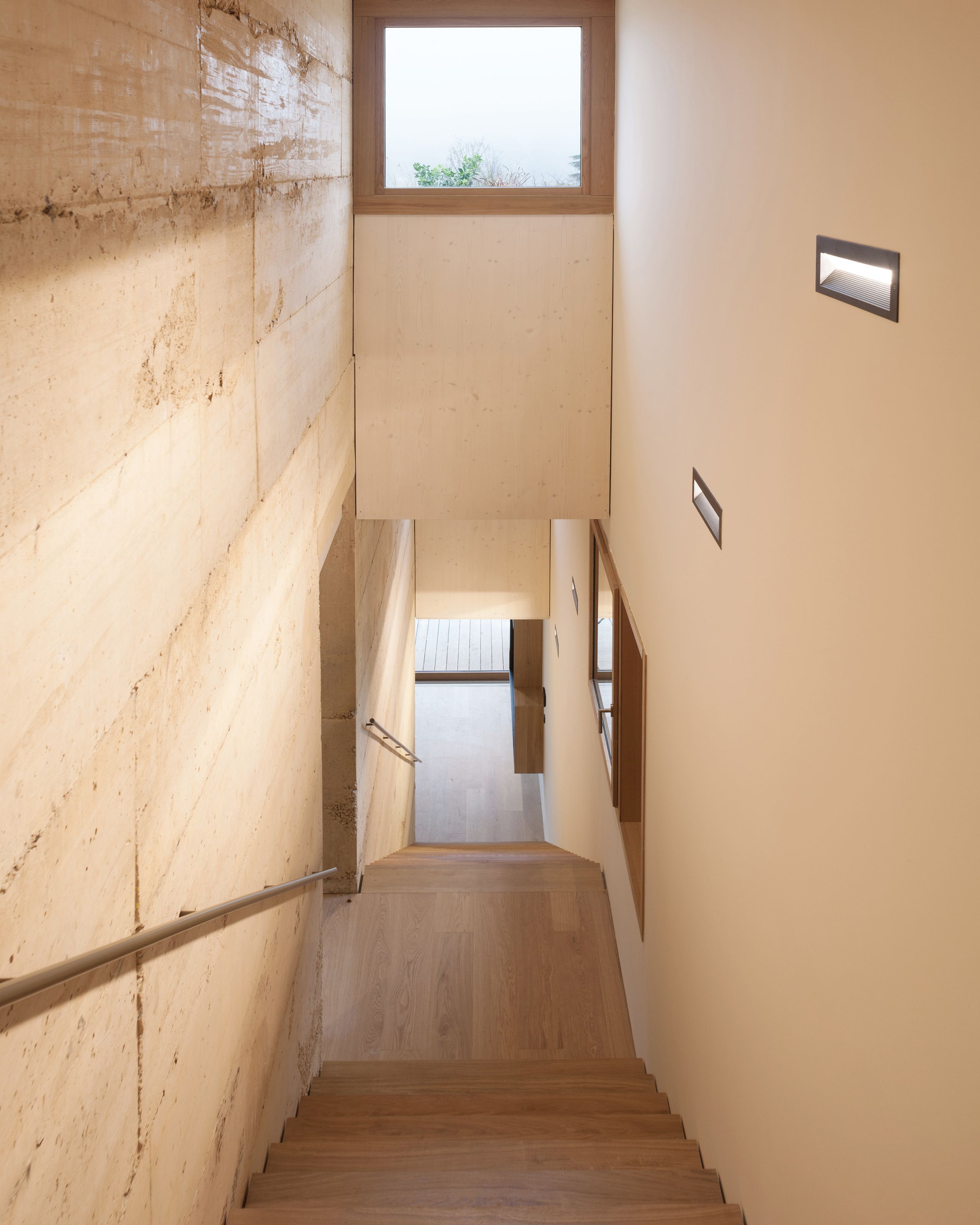 Escalier continu en bois de la maison à Saint-Cyr-au-Mont-d’Or, encadré de murs en béton teinté stratifié, éclairage mural intégré et vue cadrée sur le paysage.