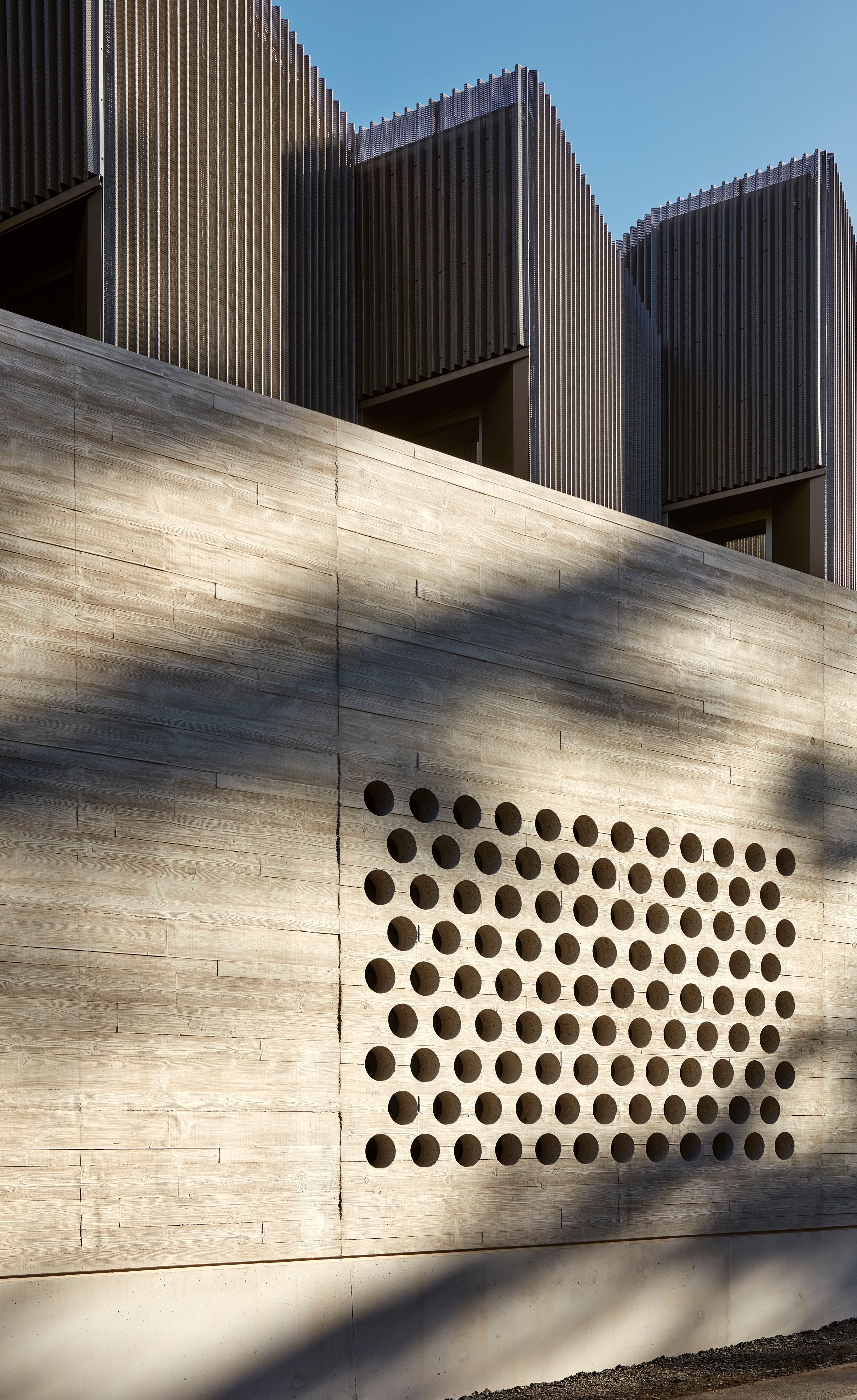 Facade detail of Maison des Arts in Charbonnières-les-Bains, showing perforated concrete base below corrugated metal-clad upper music volume in evening light.