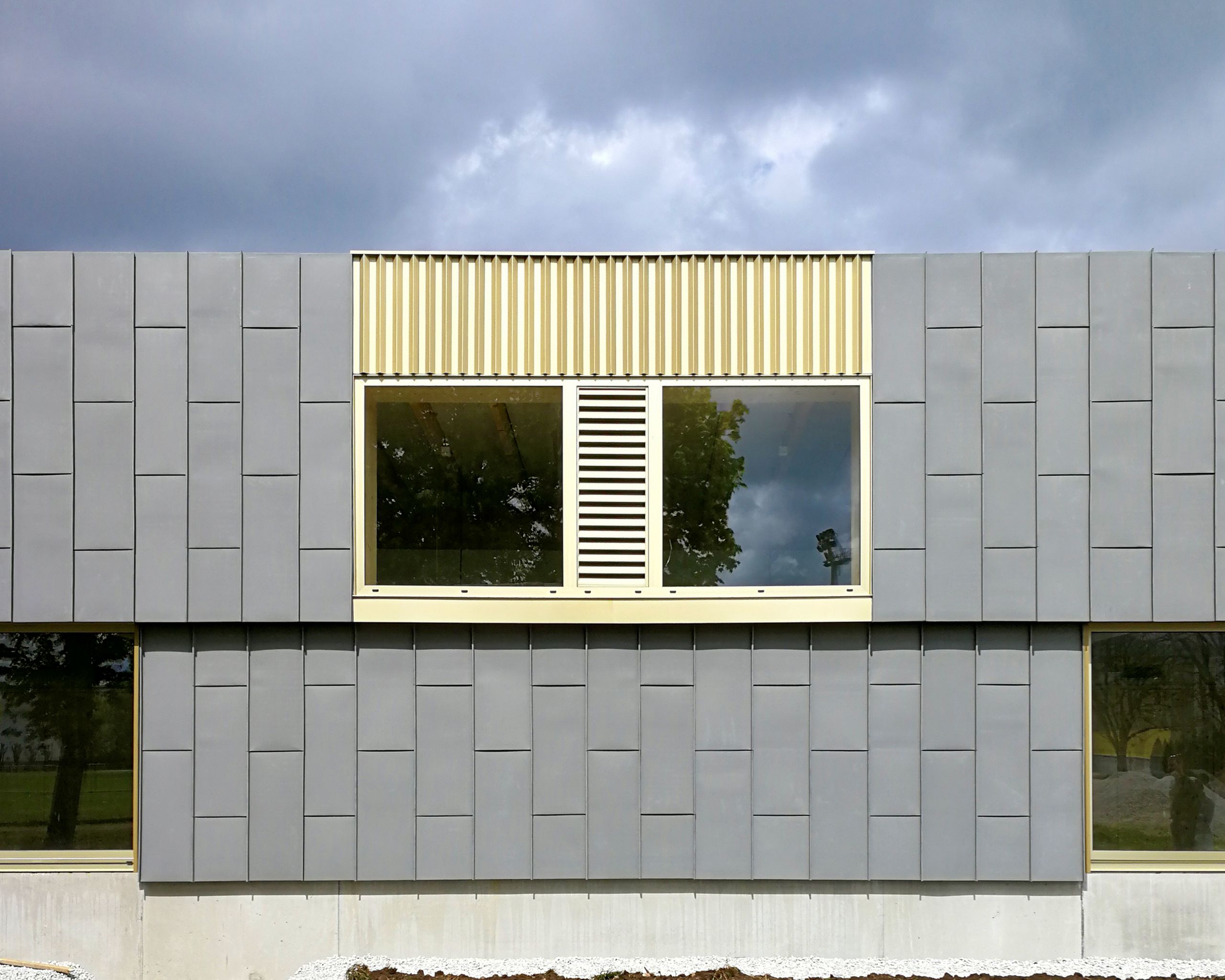 Façade de l’école élémentaire Hubert Reeves à Champagnole, bardage gris modulaire, bandeau vitré et persienné en bois clair cadrant le paysage environnant.