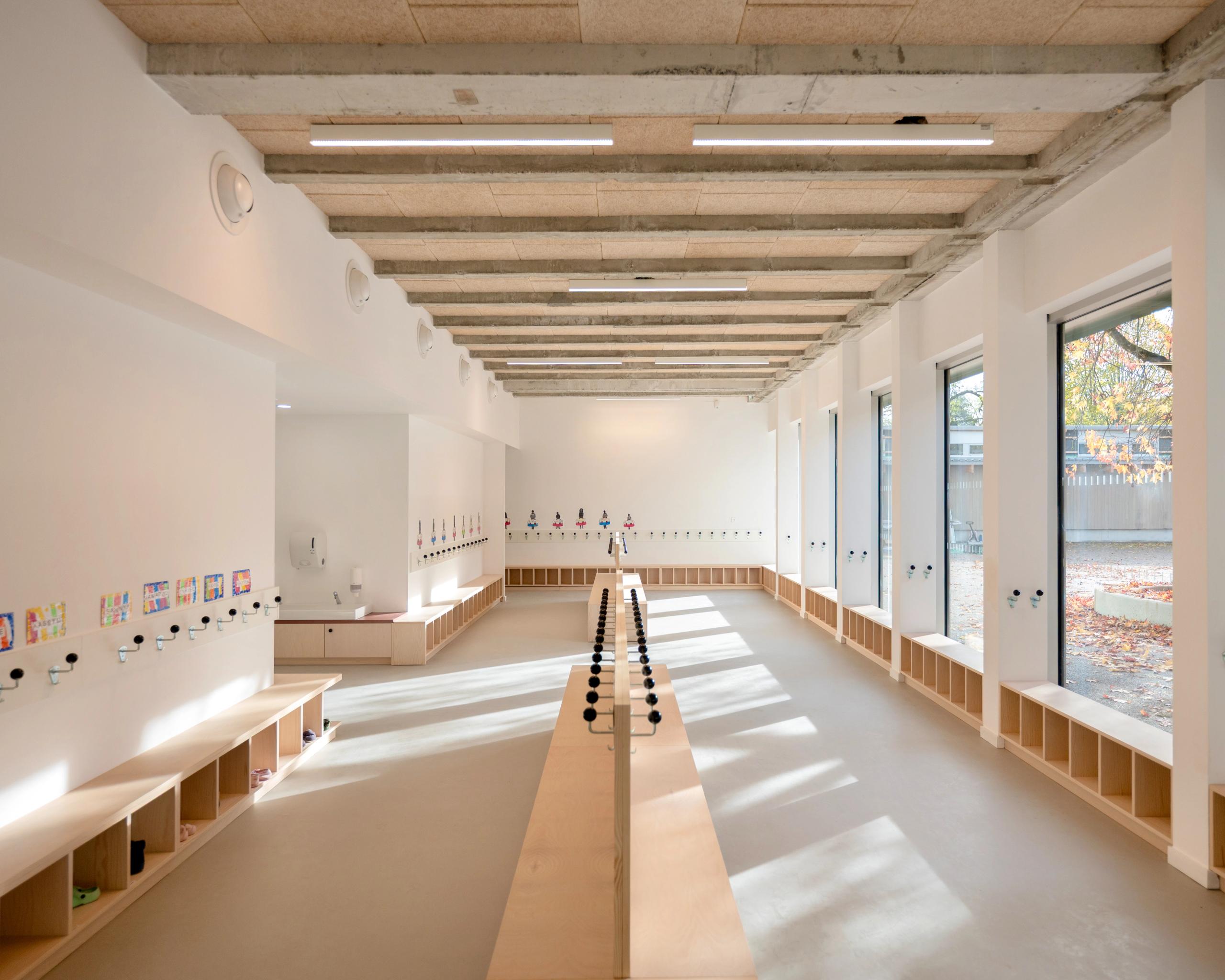 Hall du groupe scolaire du Bouchet à Bonneville : espace traversant lumineux, structure béton apparente, menuiseries bois intégrées et grandes baies vitrées sur cour.