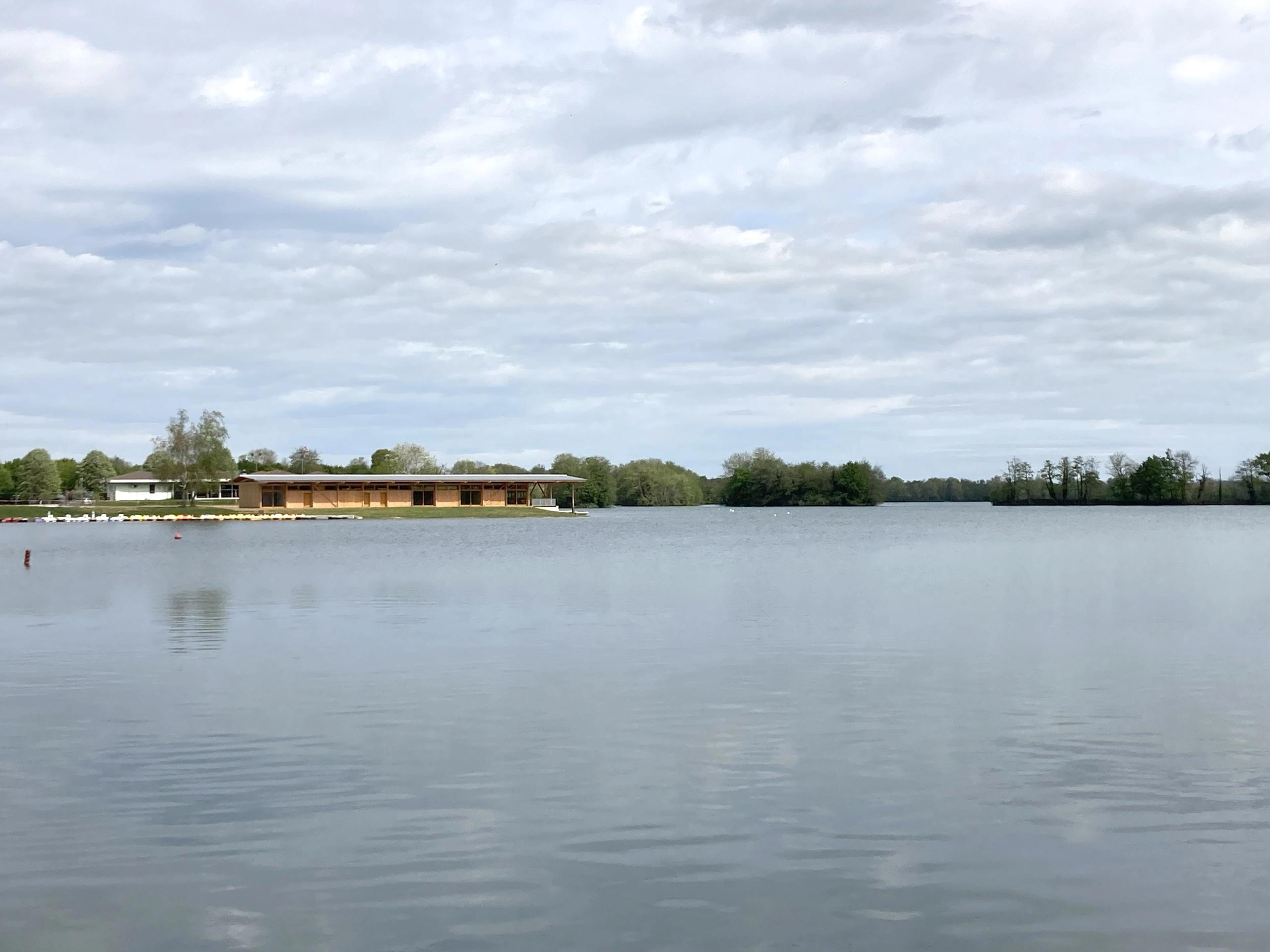 Vue lointaine de la Maison des sports à Malafretaz, volume bas en bois et toiture débordante en rive de lac, inséré dans un paysage arboré et ouvert.