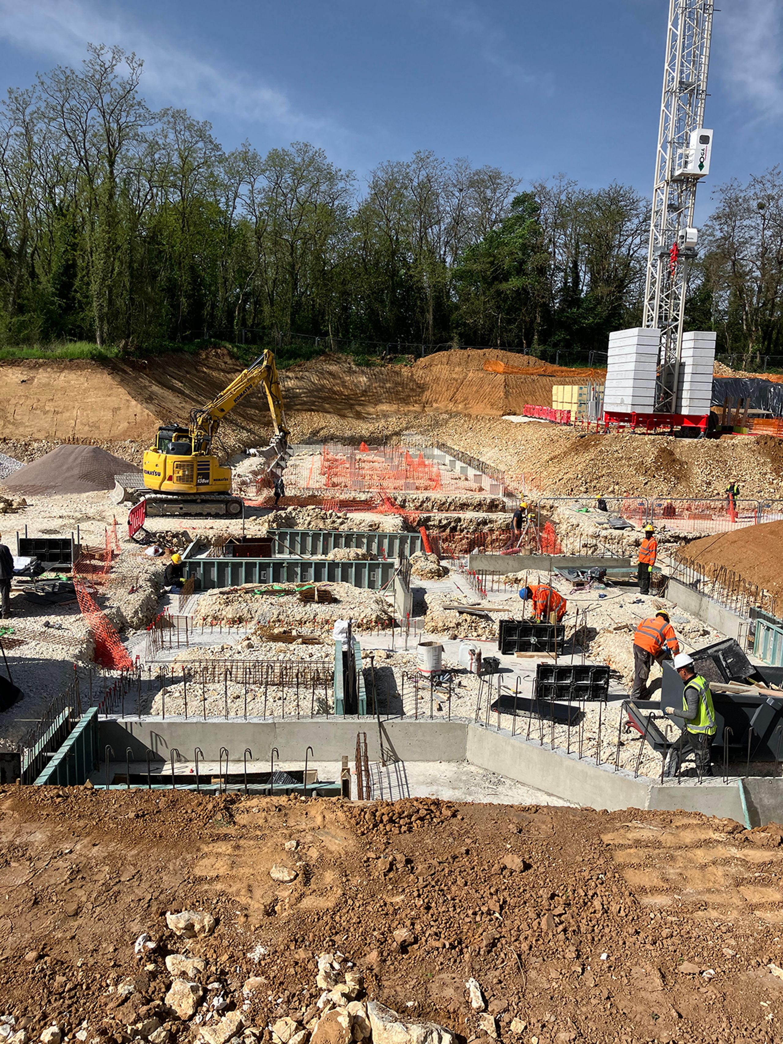 Construction site of the Auxerre nursing home, showing excavated terraces, concrete foundations, crane and earthmoving equipment against a wooded backdrop.