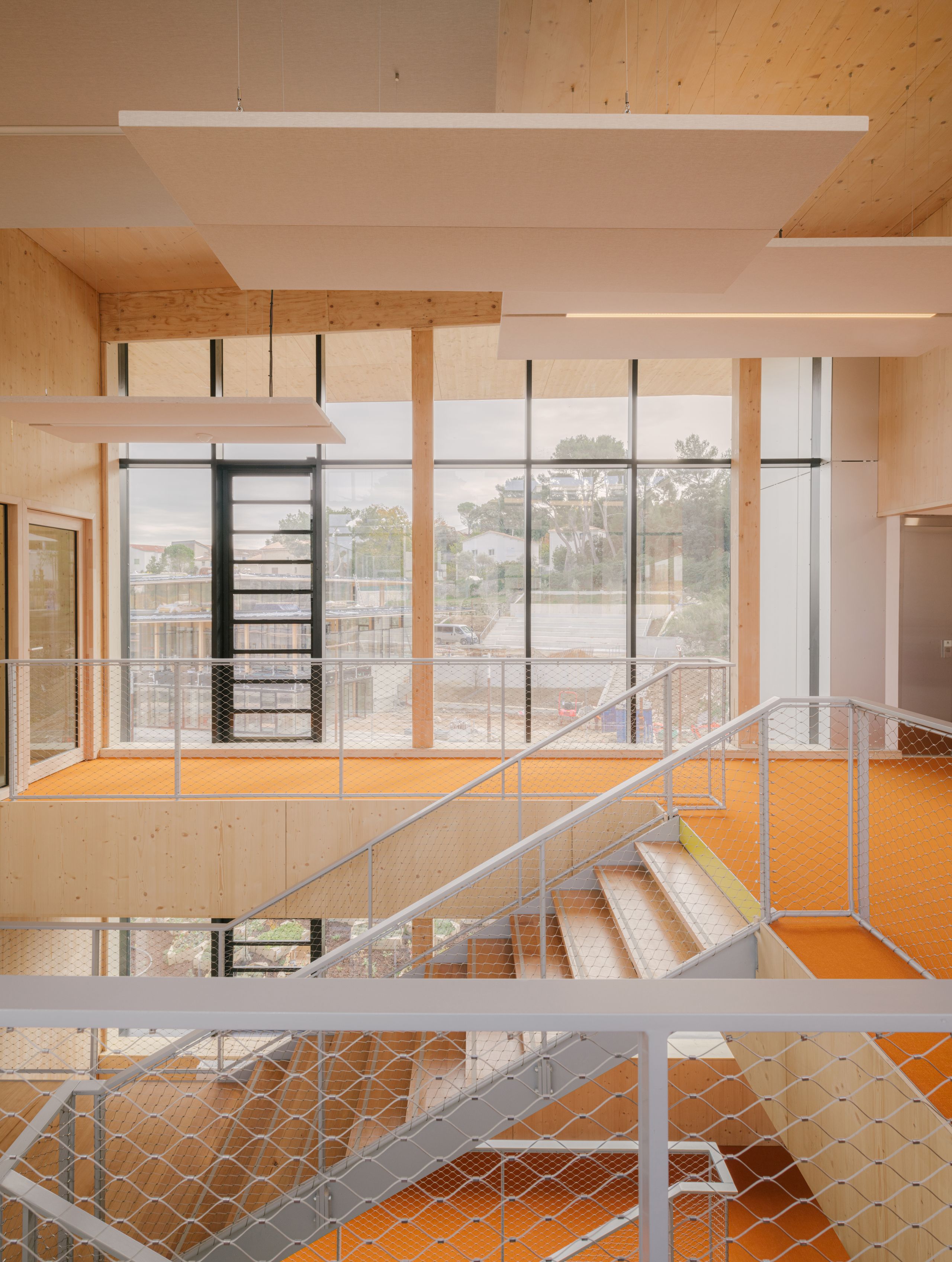 Escalier central en bois et métal du groupe scolaire Claudie Haigneré aux Pennes-Mirabeau, baigné de lumière naturelle par une grande façade vitrée.