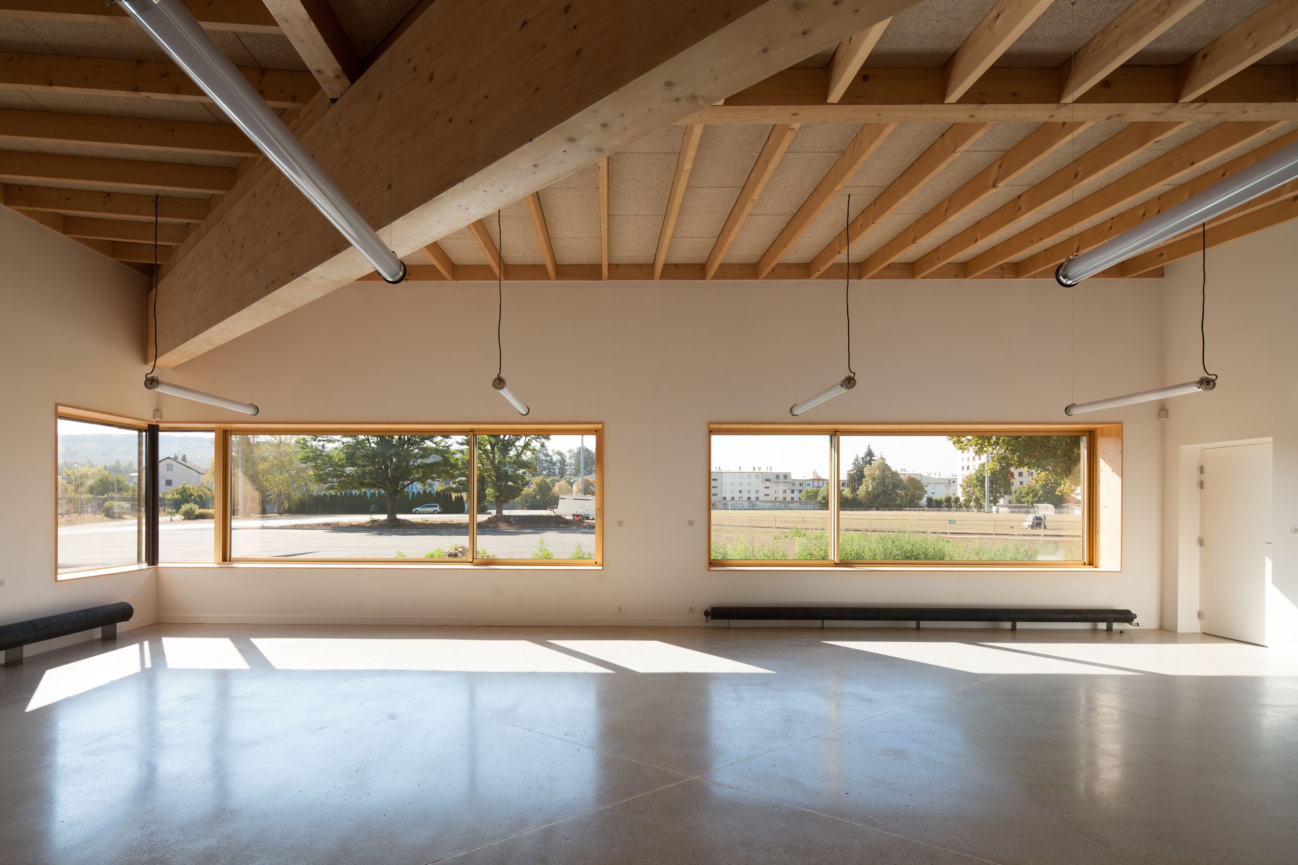 Intérieur de l’école élémentaire Hubert Reeves à Champagnole : grande salle vitrée, ossature bois apparente, dalles béton et vues dégagées sur le paysage périurbain.
