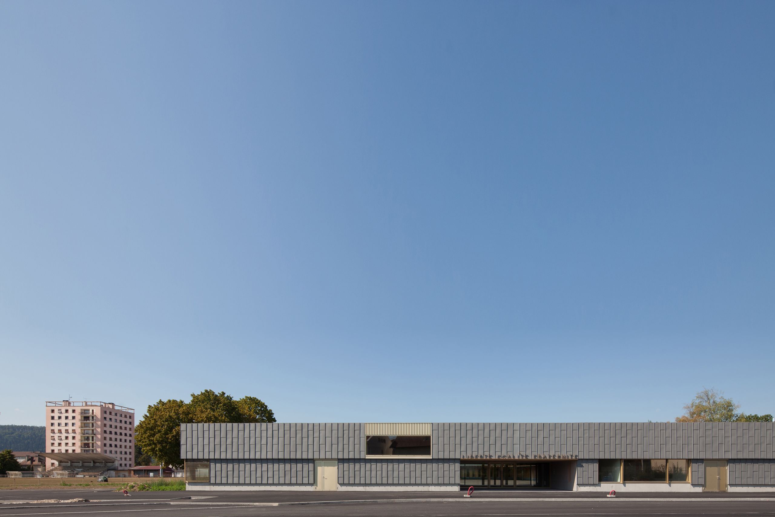 Façade principale de l’école élémentaire Hubert Reeves à Champagnole, volume bas en zinc à trame régulière, percé de baies cadrant le paysage périurbain.