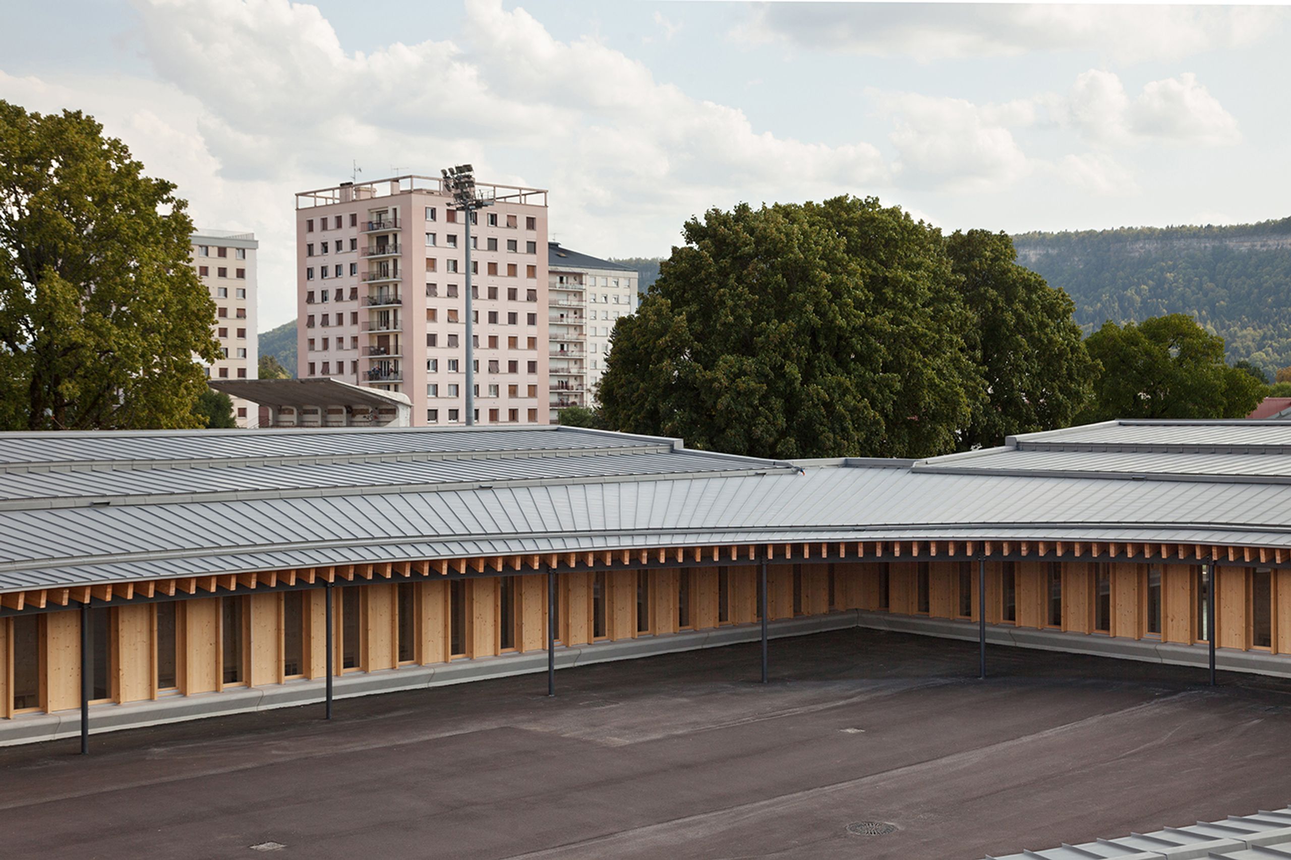 Vue de la cour elliptique de l’école élémentaire Hubert Reeves à Champagnole, toiture en zinc en impluvium et façades à ossature bois ouvertes sur le paysage.