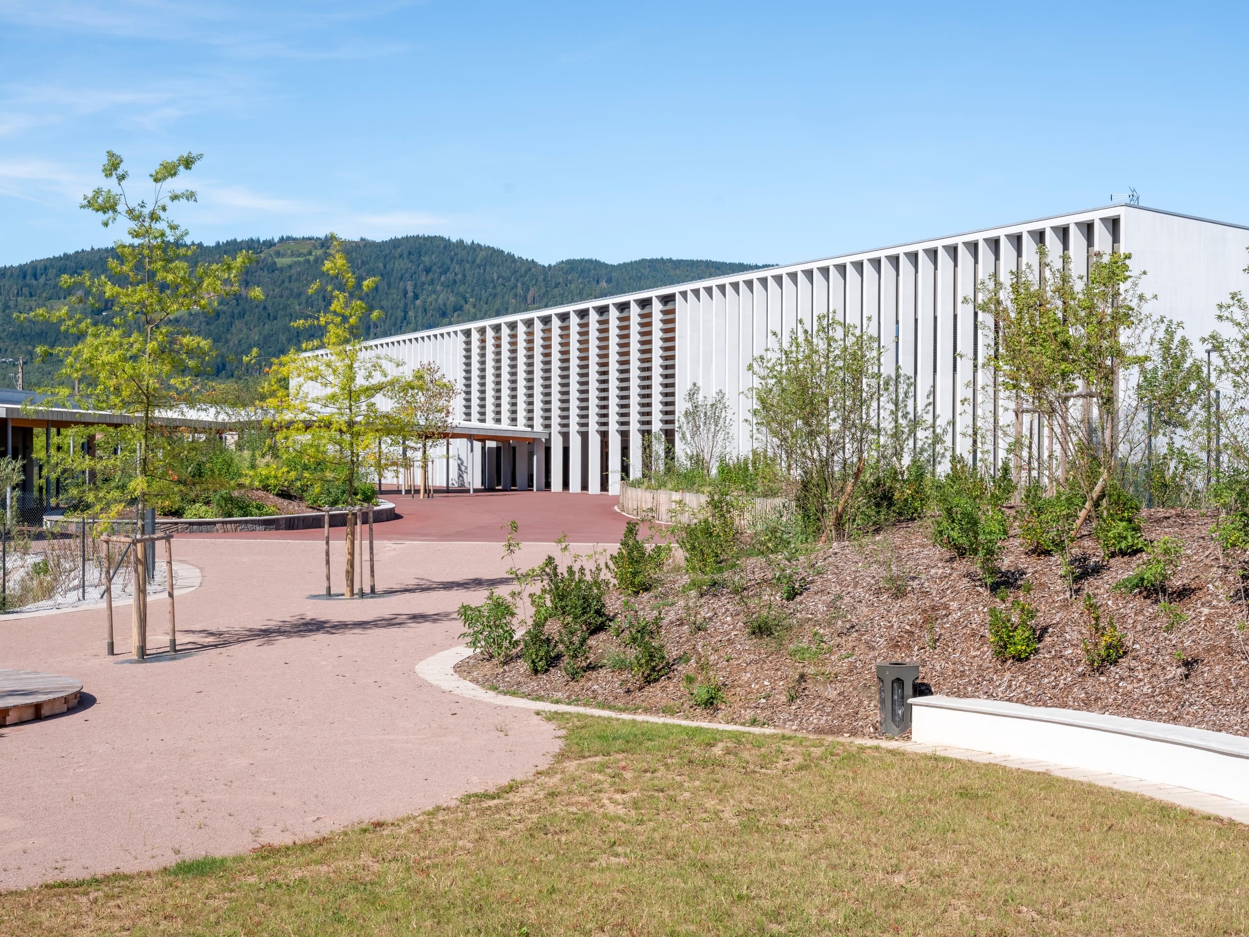 Vue du Collège du Ban à Vagney : volume linéaire à portique rythmé, façades blanches, préau ouvert sur cour minérale et aménagements paysagers en lisière de montagne.