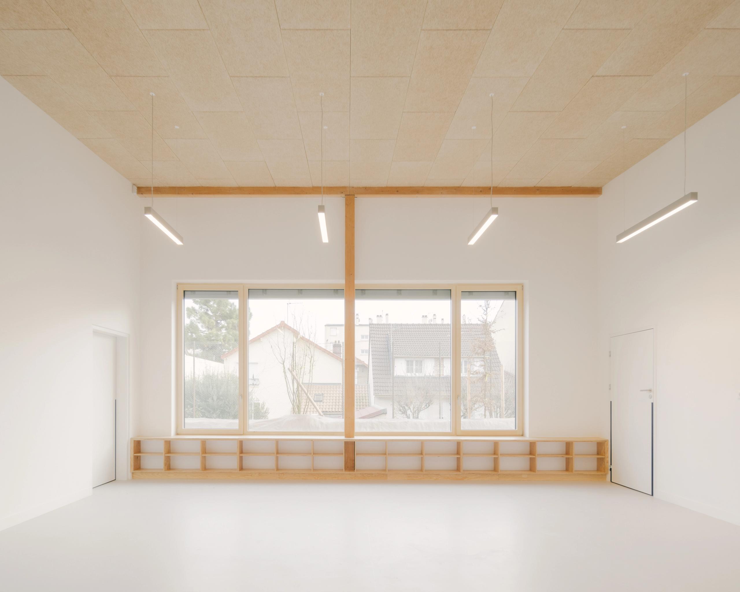 Intérieur lumineux d’une salle des groupes scolaires Jacqueline Auriol et Jean Jaurès au Bourget, avec ossature bois apparente, plafond acoustique et grande baie vitrée sur le quartier.