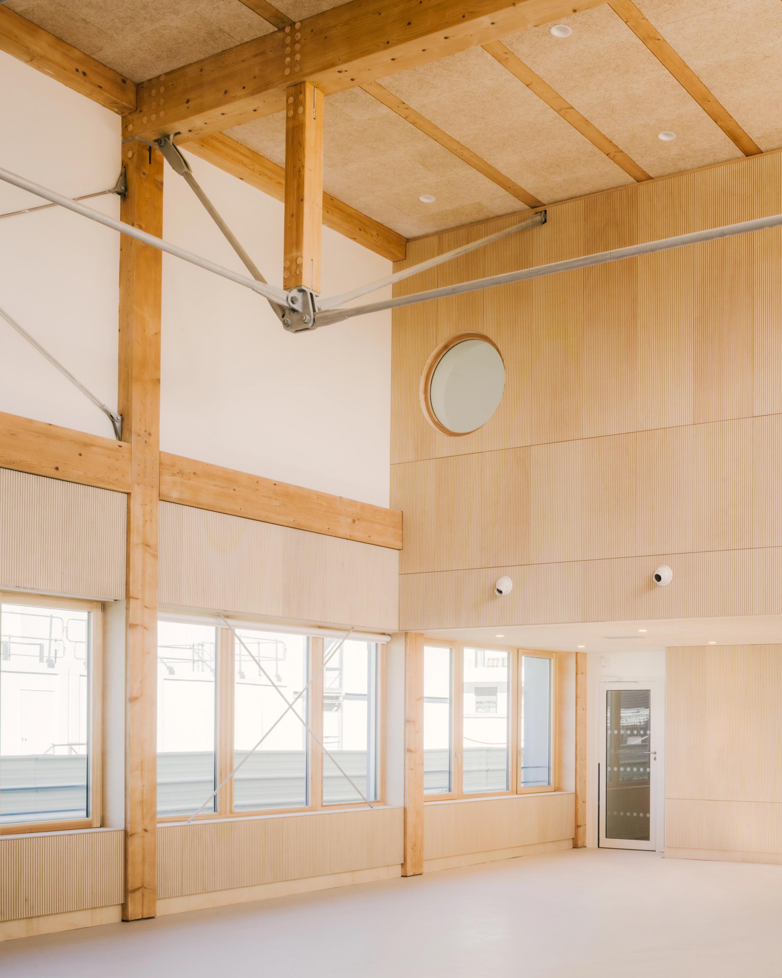 Intérieur du groupe scolaire Dominique Frelaut à Colombes : grand volume en structure bois apparente, bardage intérieur clair, baies vitrées et porte vitrée sur cour.