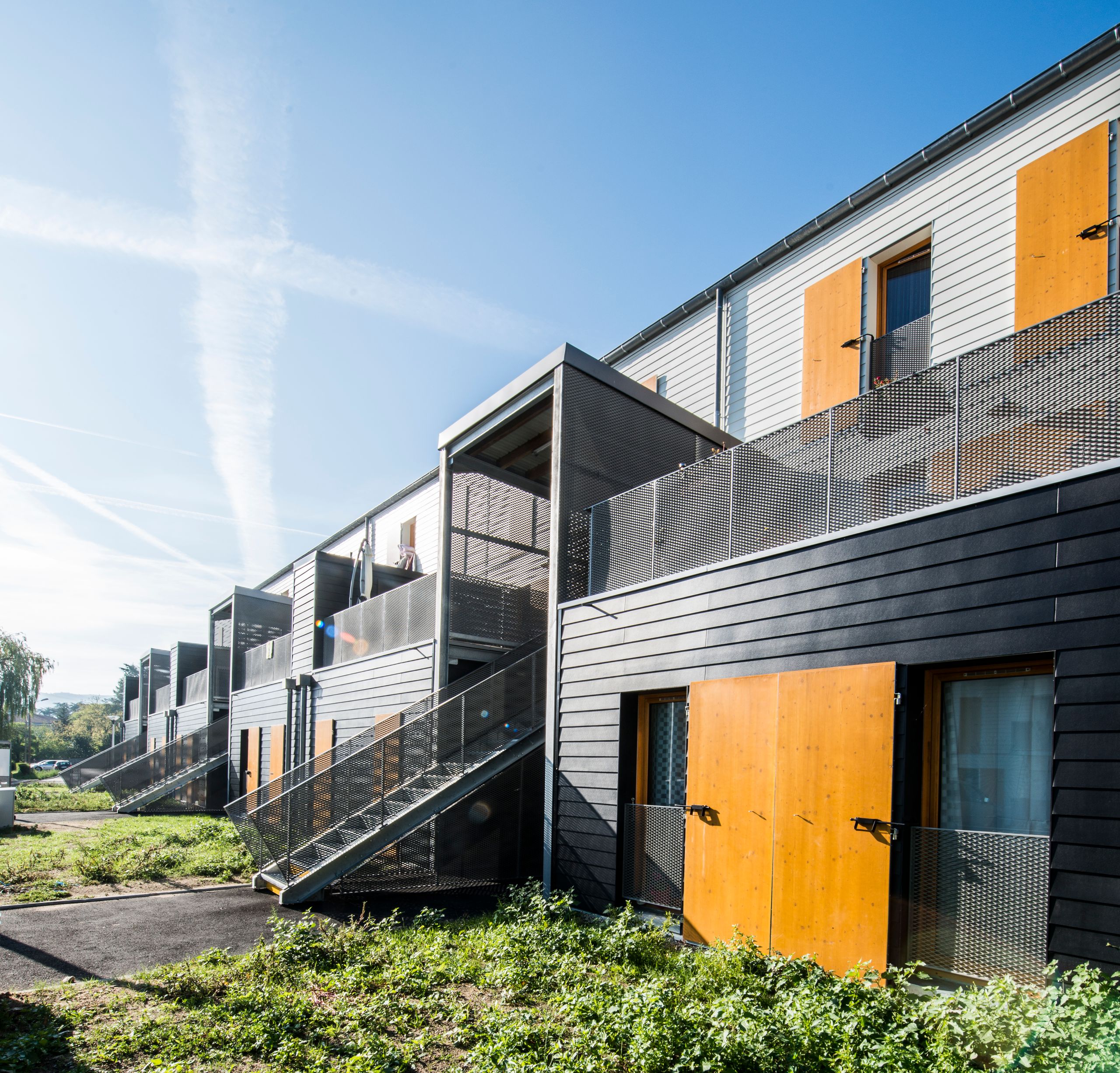 Vue des 60 logements à Rive-de-Gier : volumes fragmentés en terrasses, structure 5D apparente, bardage bois et métal, escaliers extérieurs sur site en pente.