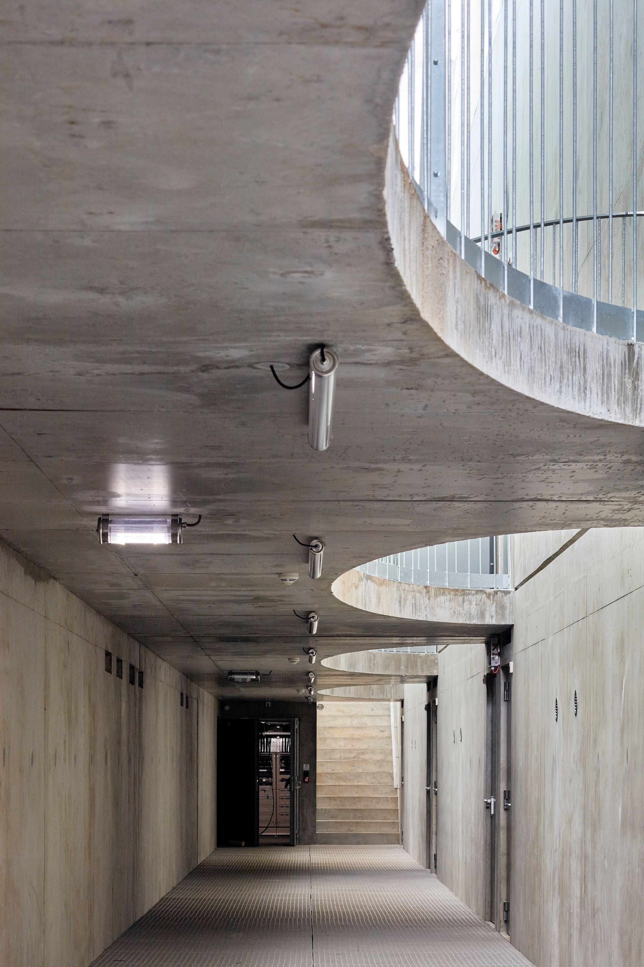 Circulation en triple hauteur du poste source de Cusset à Villeurbanne, couloir en béton brut éclairé par des percements circulaires et des luminaires techniques.