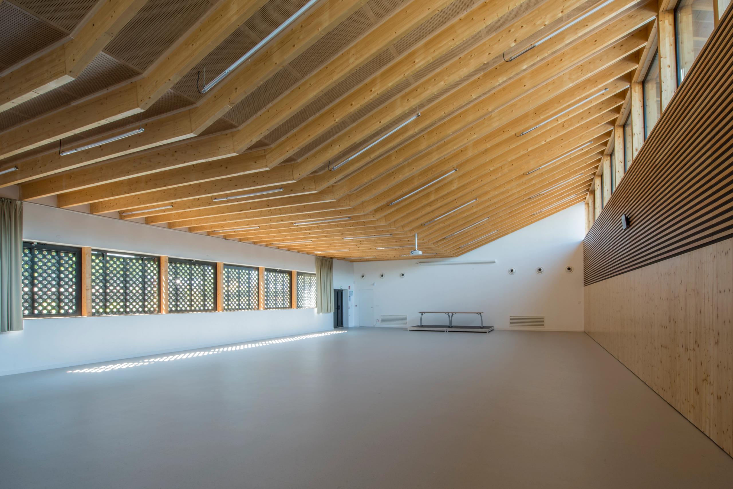 Grande salle du collège Pierre-Joseph Proudhon à Besançon, charpente bois apparente, façades vitrées filtrantes et parois claires, éclairage naturel latéral.