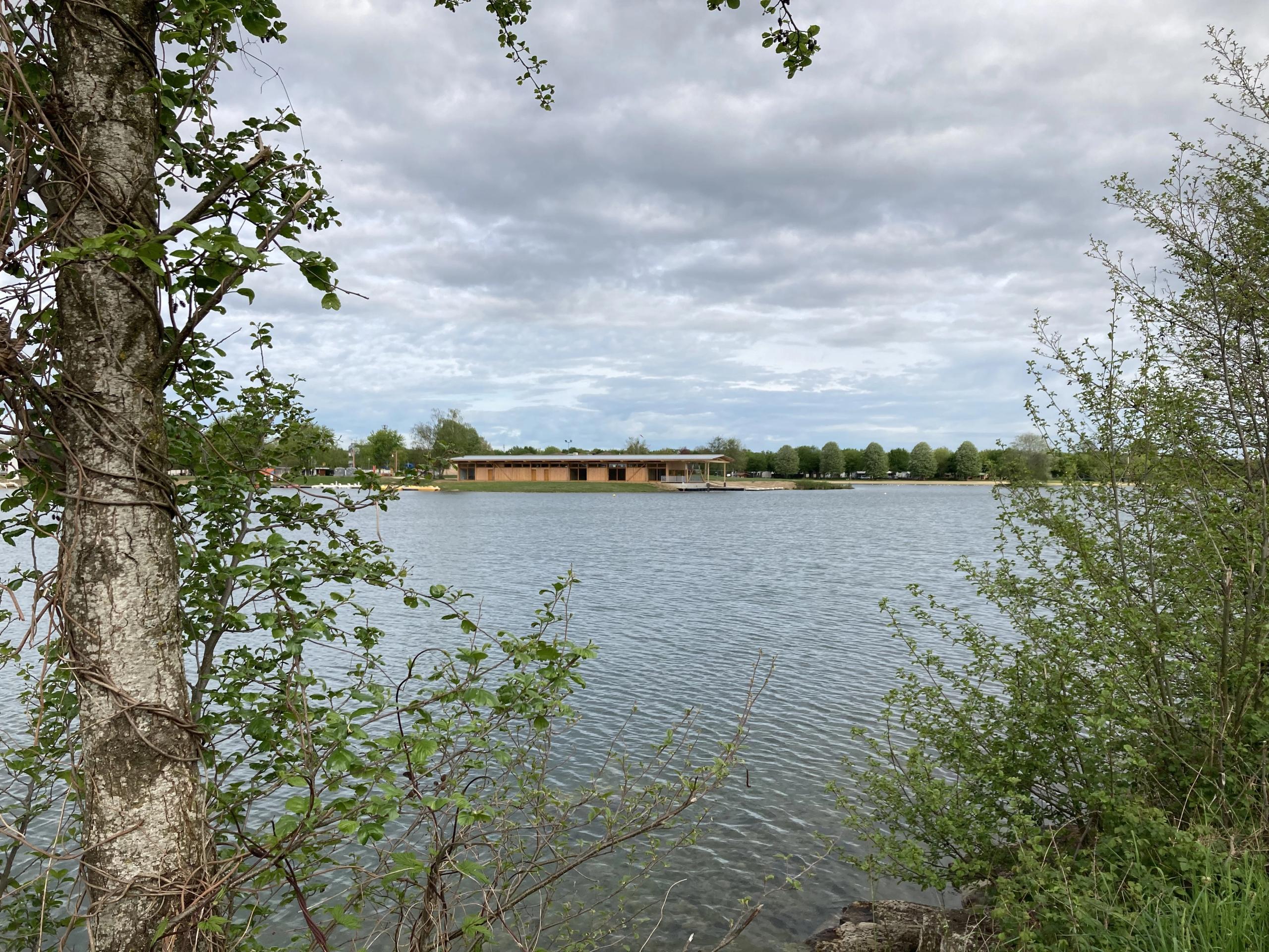 Maison des sports à Malafretaz en rive de plan d’eau, volume bas en bois et toiture plate, largement vitré, inséré dans un paysage arboré et ouvert.