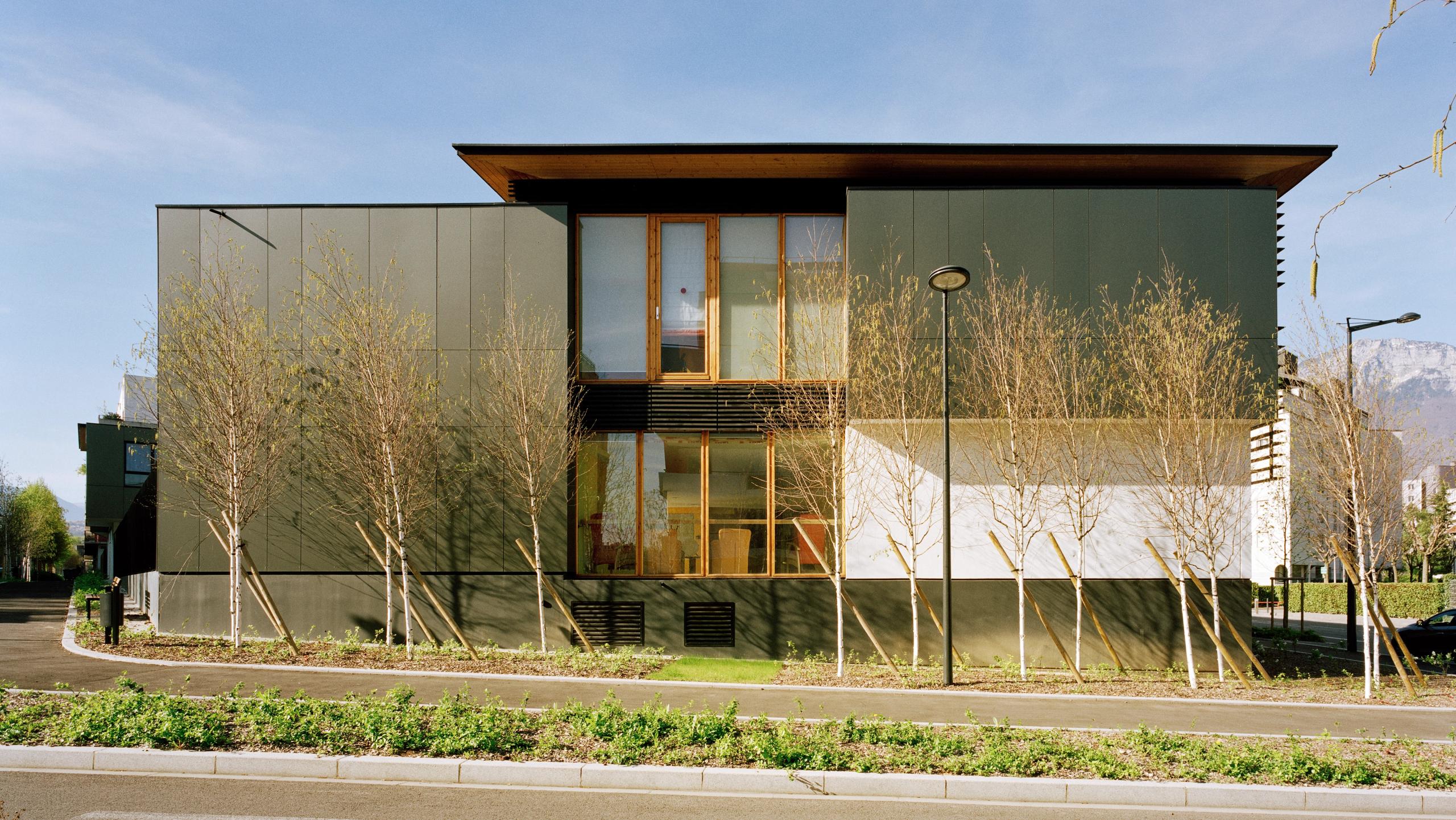 Façade de l’EHPAD à Grenoble par Tectoniques, volume compact à deux niveaux, bardage sombre, grandes baies bois, toiture débordante et jardins plantés en pied de bâtiment.