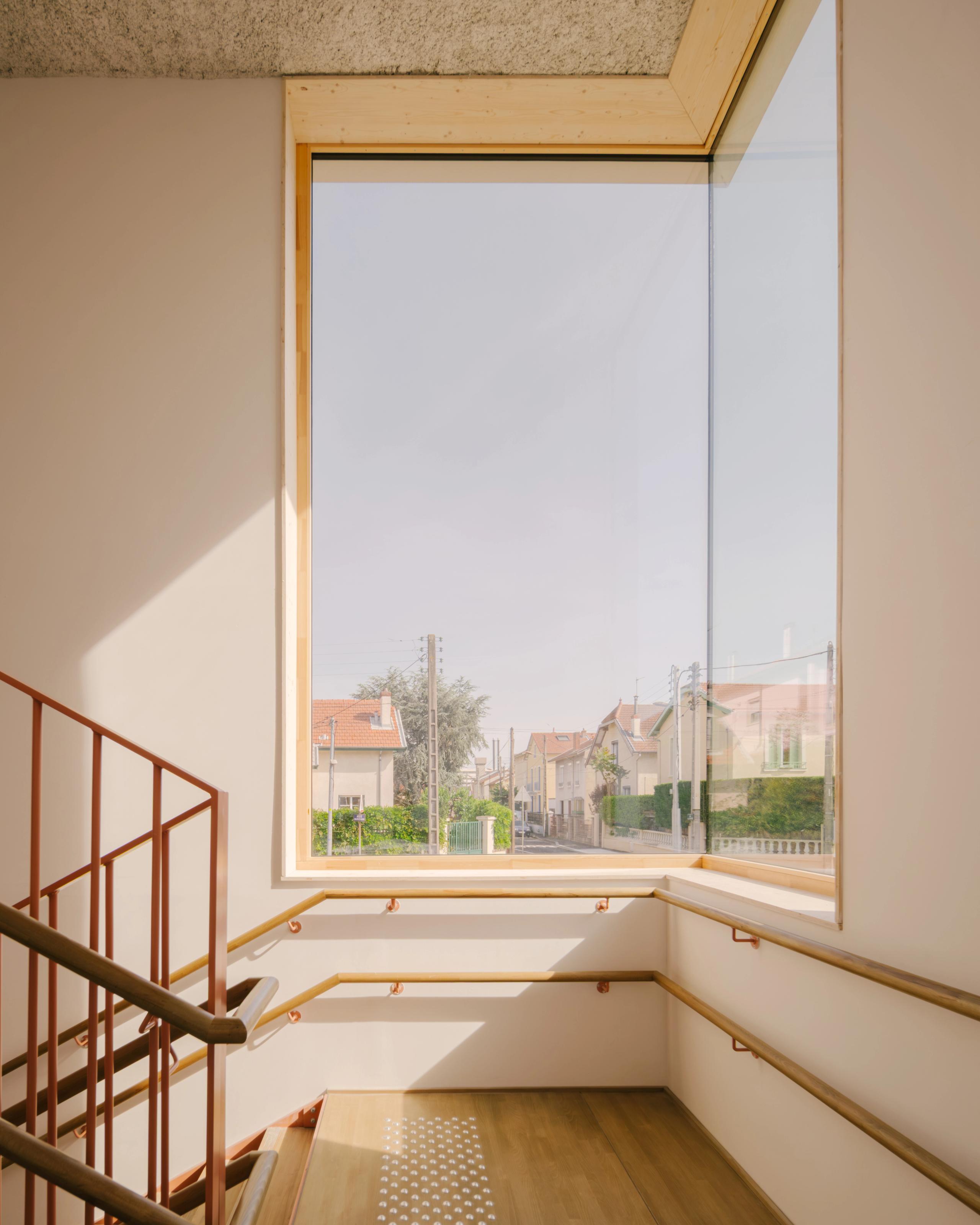 Escalier du groupe scolaire Kennedy à Lyon, éclairé par une grande baie vitrée bois, avec garde-corps métalliques et mains courantes continues sur plancher bois.