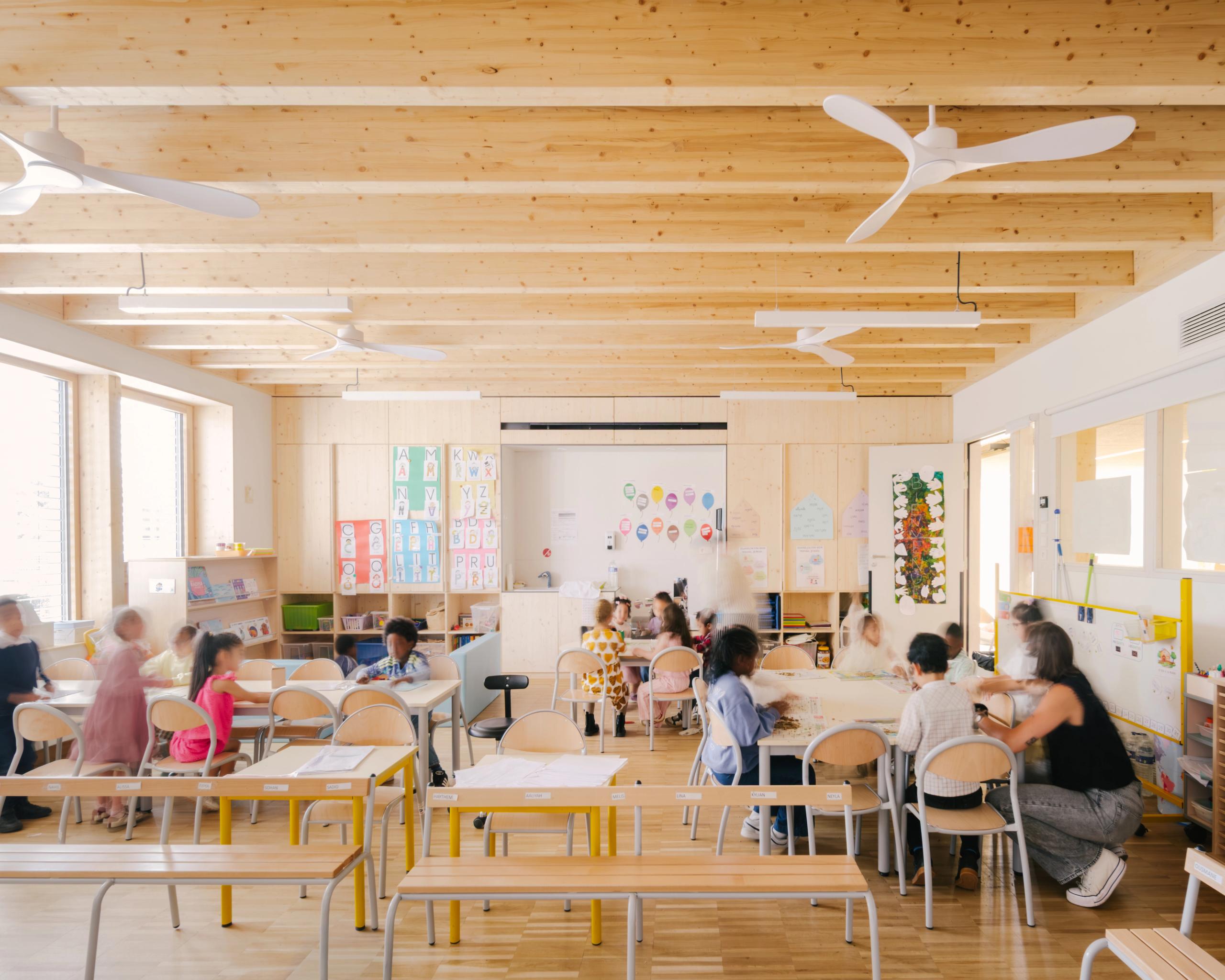 Salle de classe du groupe scolaire Kennedy à Lyon, structure bois apparente, lumière naturelle latérale, mobilier clair modulable et plafond à poutres avec ventilateurs.