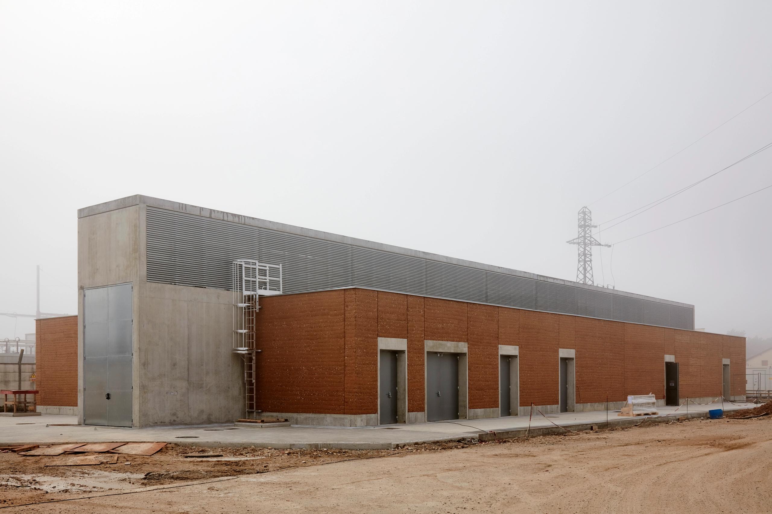 Poste source de Cusset à Villeurbanne : volume industriel linéaire en béton brut, pisé et acier, façades rythmées de portes techniques sous ciel brumeux.