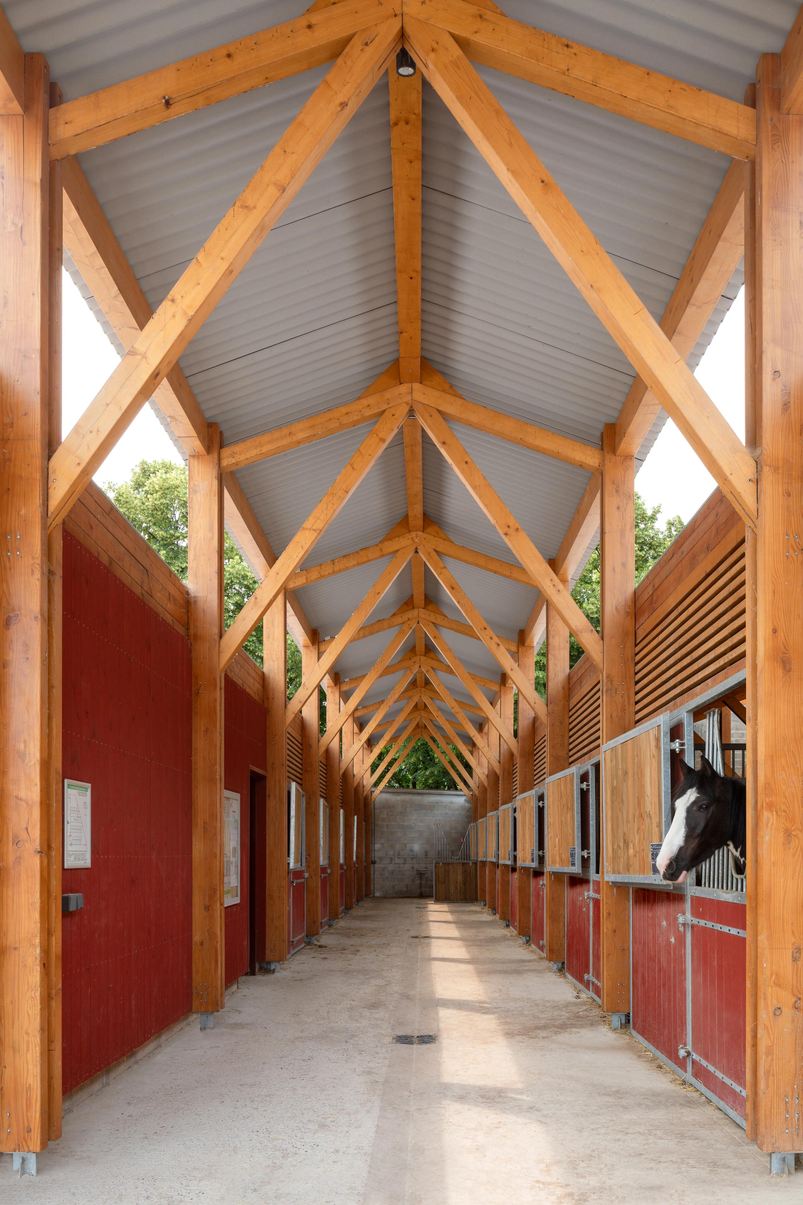 Timber-framed stable corridor at the Gennevilliers equestrian center, with Falun red cladding, metal roof, and horses in open stalls along one side.