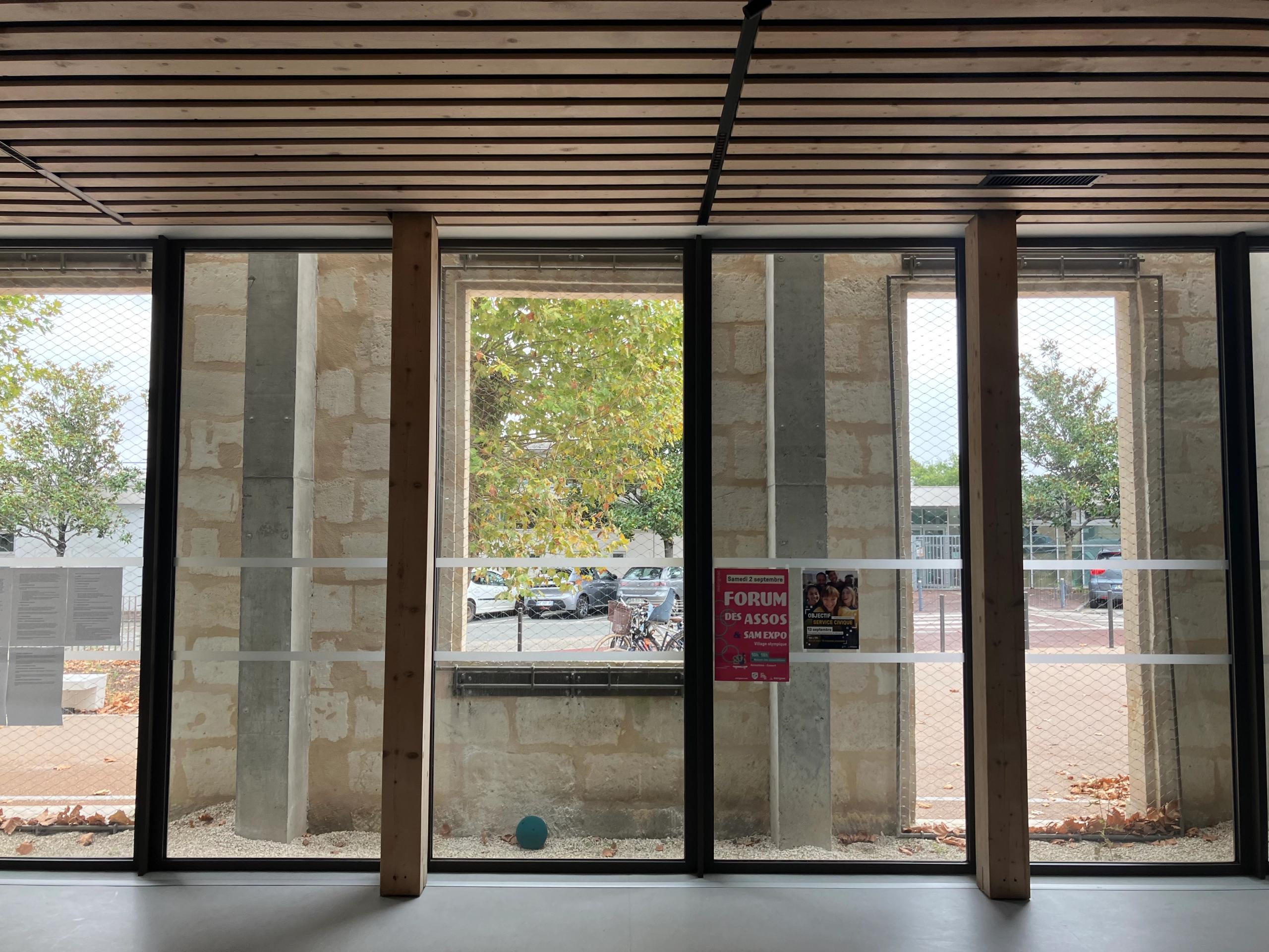Intérieur de la maison des habitants à Mérignac : façade existante en pierre percée de grandes baies vitrées, structure bois apparente et vue sur l’espace public arboré.