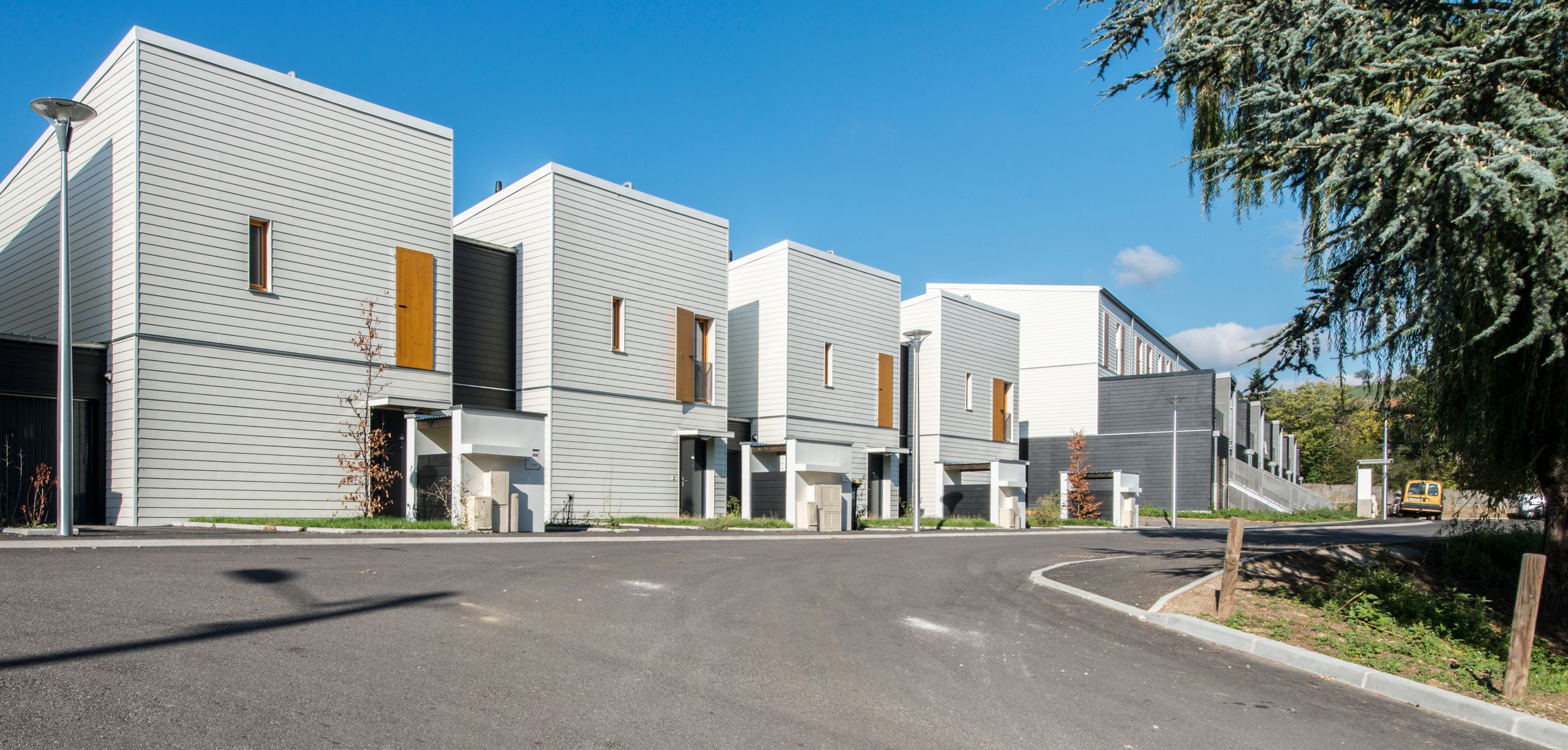 Vue de la livraison des 60 logements Tectoniques à Rive-de-Gier : volumes fragmentés en bande, façades à bardage horizontal clair et modules 5D sur voie courbe.