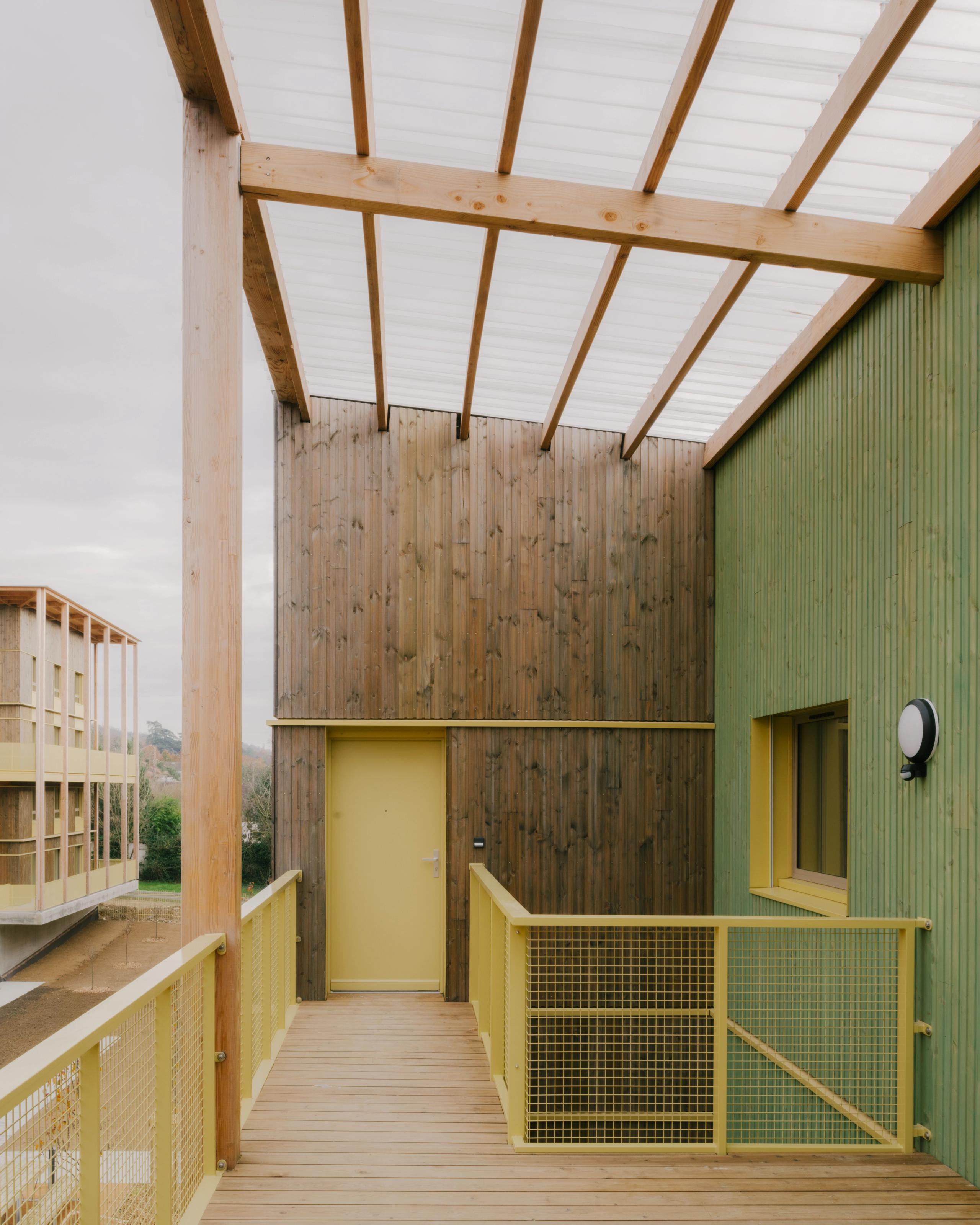Coursive extérieure des 84 logements à Trévoux par Tectoniques, structure bois, garde-corps métalliques jaunes et façades bois et bardage vert sous auvent translucide.