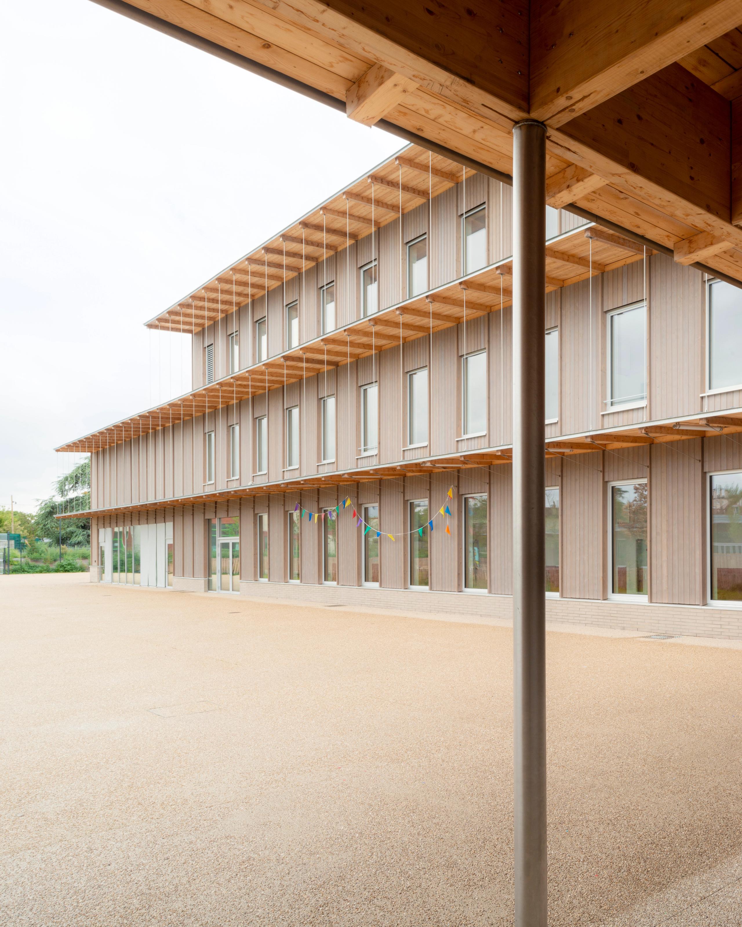 Façade bois des groupes scolaires Jacqueline Auriol et Jean Jaurès au Bourget, sous larges débords de toiture en bois, ouvrant sur une vaste cour minérale.