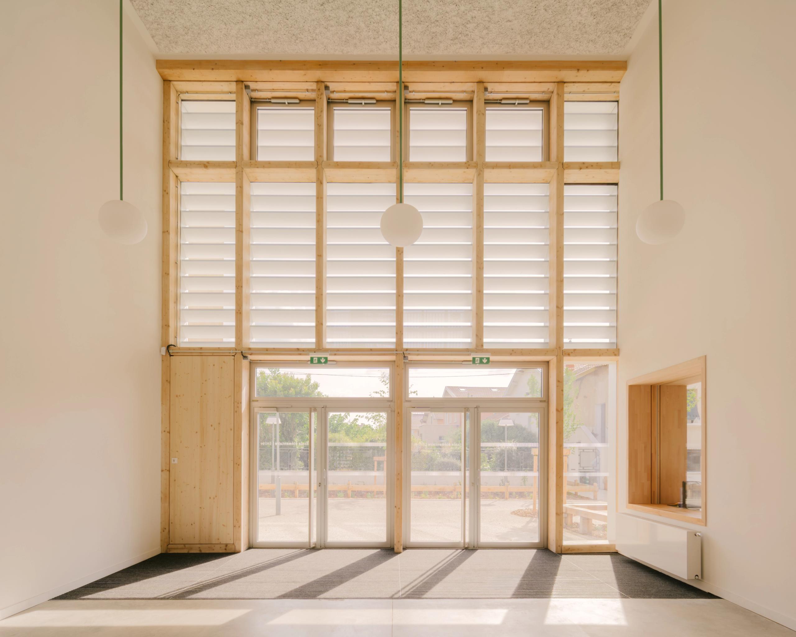 Hall traversant du groupe scolaire Kennedy à Lyon, double hauteur en structure bois, grande façade vitrée à lames orientables ouvrant sur la cour végétalisée.