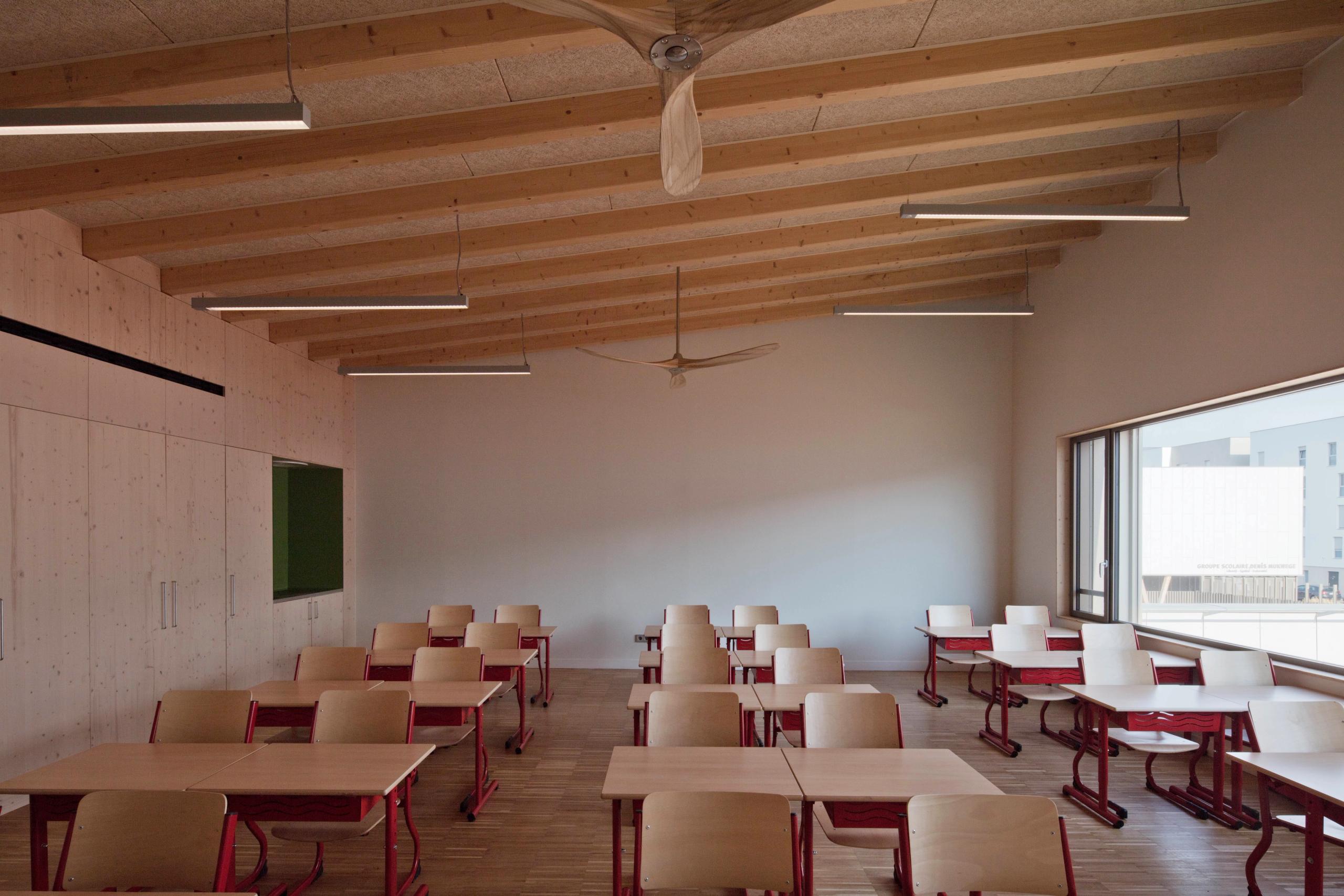 Salle de classe du groupe scolaire Denis Mukwege à Melun, structure bois apparente, mobilier rouge modulable, grande baie vitrée et éclairage linéaire.