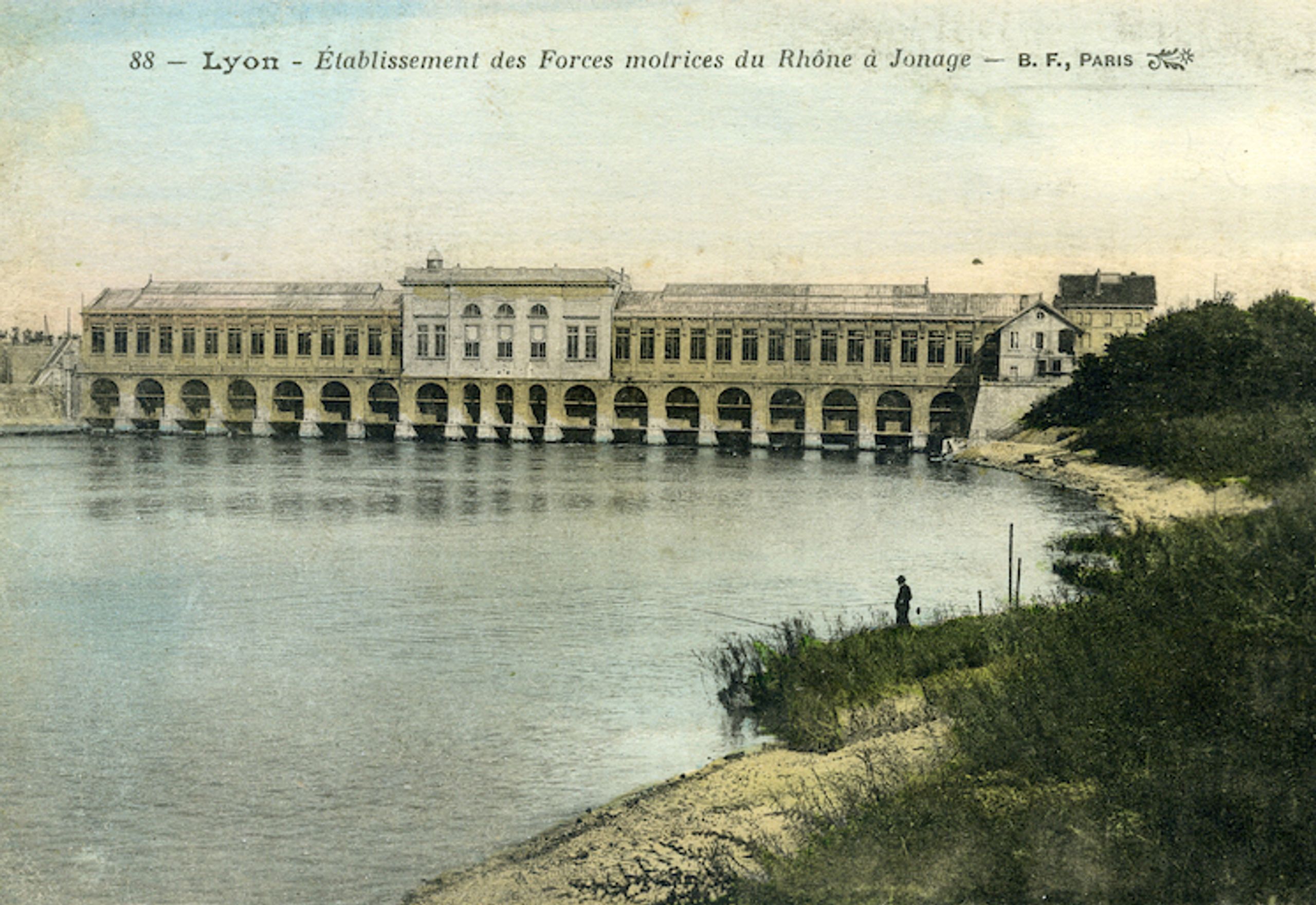 Vue ancienne de la centrale hydroélectrique de Cusset à Villeurbanne, long bâtiment industriel rythmé d’arcades sur le Rhône, inscrit dans un paysage fluvial.