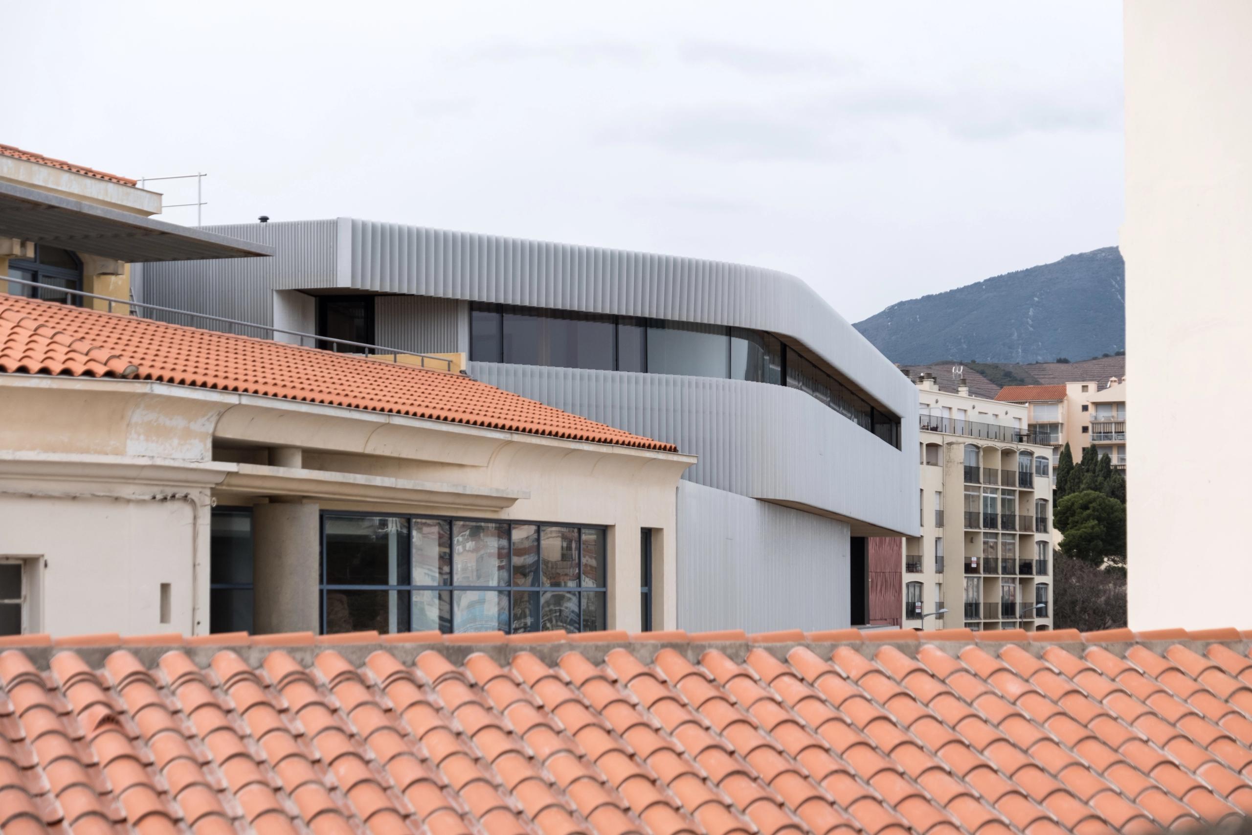 Vue du Biodiversarium à Banyuls-sur-Mer : volume compact en bardage métallique ondulé gris, percé de longues baies vitrées, inséré dans le tissu urbain côtier.