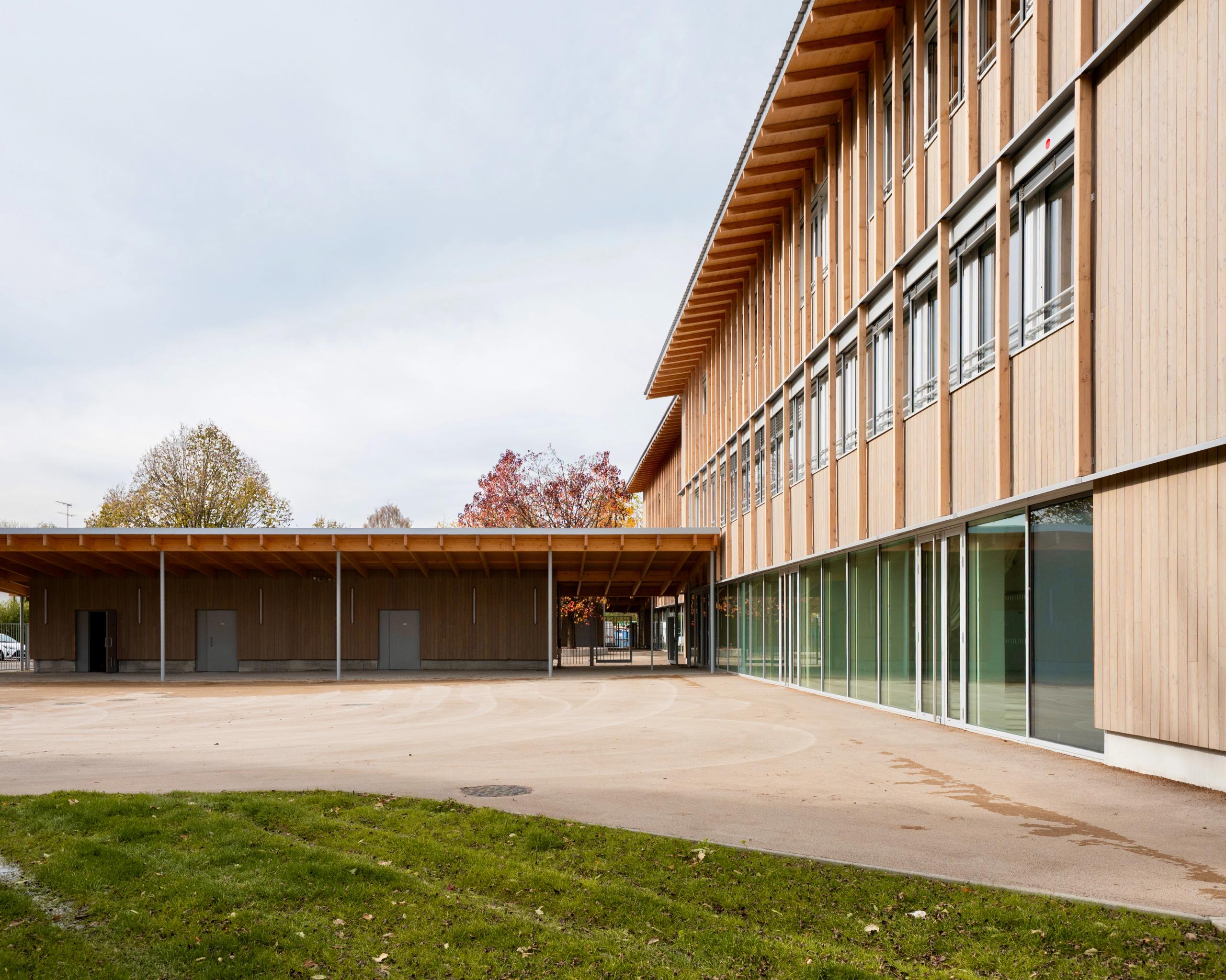Cour du groupe scolaire du Bouchet à Bonneville, façades bois surélevées, préau perpendiculaire couvert, grandes baies vitrées ouvrant sur la cour minérale.